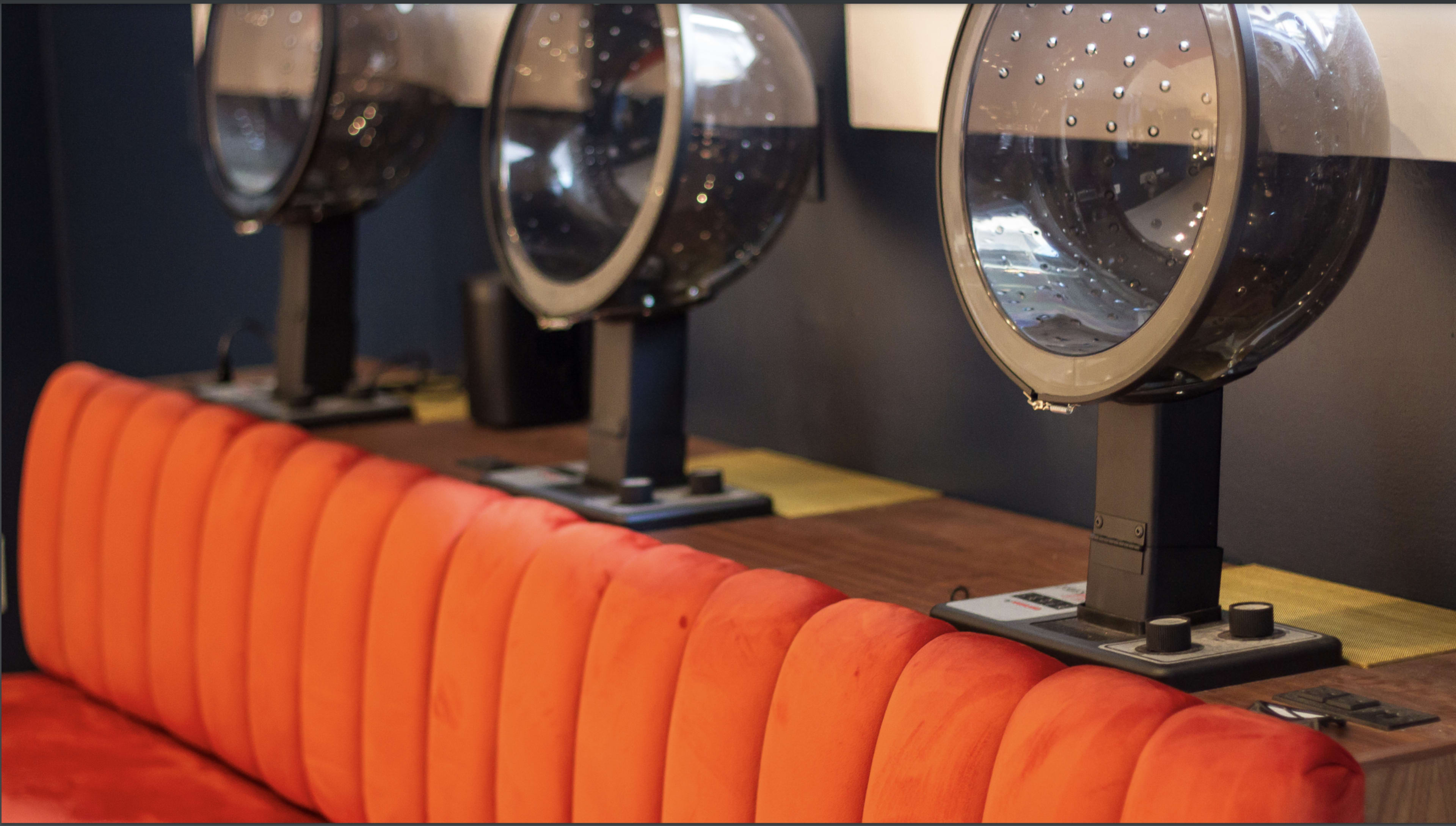 Three hairdryers on stands are positioned on a wooden counter behind an orange tufted couch.