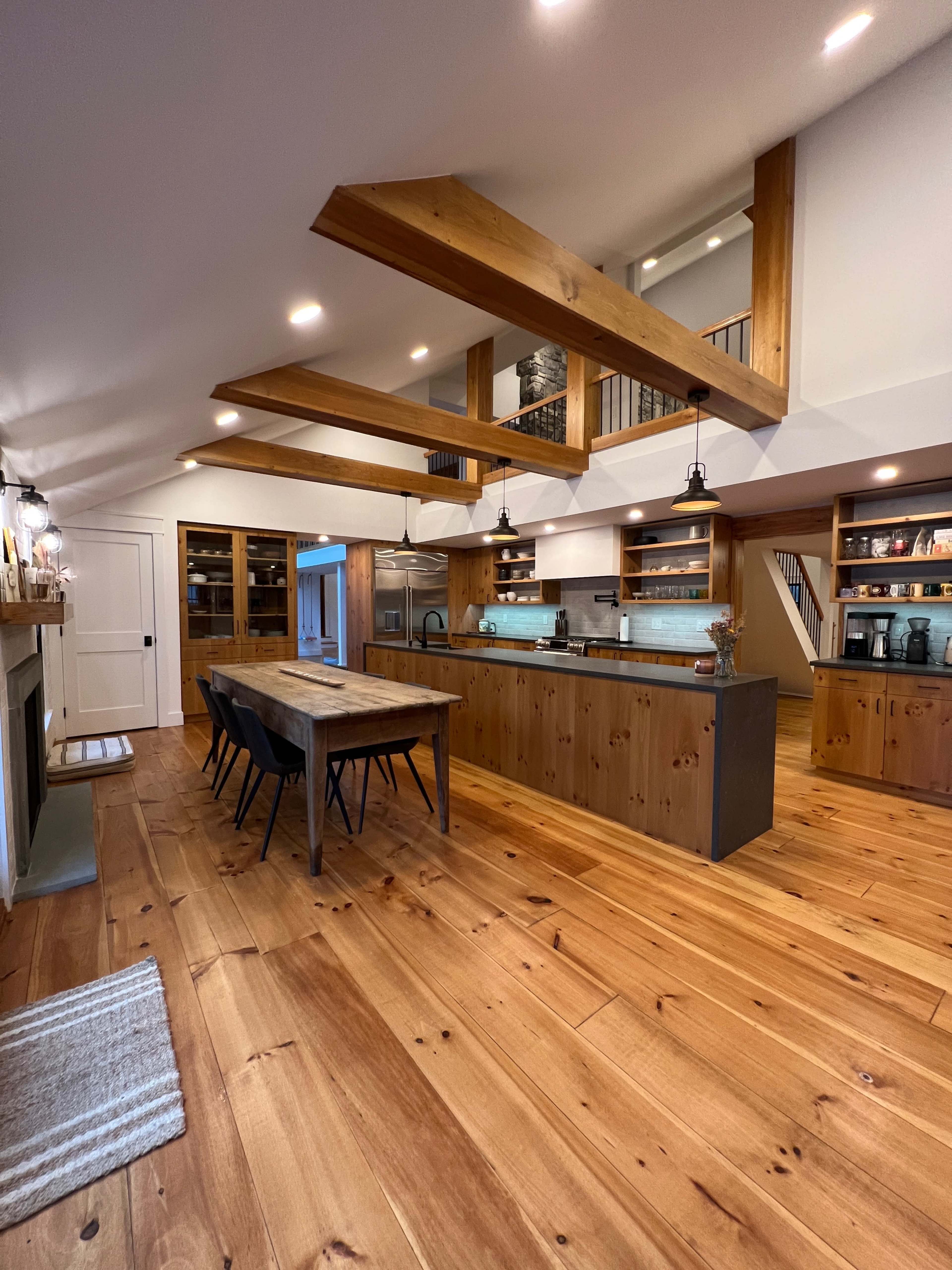 A modern kitchen featuring a large wooden island, a dining table with black chairs, and wooden beams supporting the ceiling.