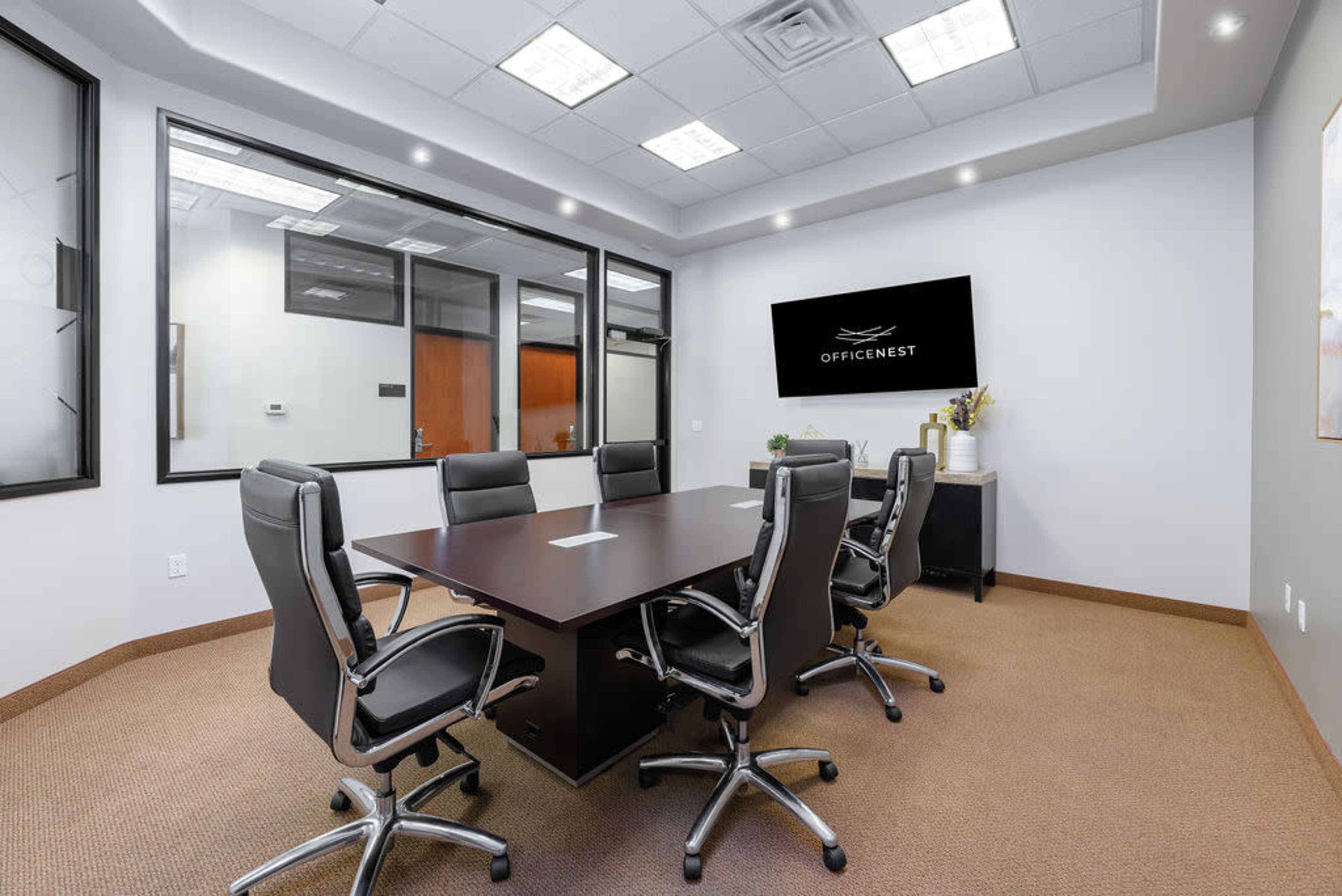A modern conference room features a large dark table surrounded by six black ergonomic chairs, with a wall-mounted television displaying the logo "OFFICENEST."