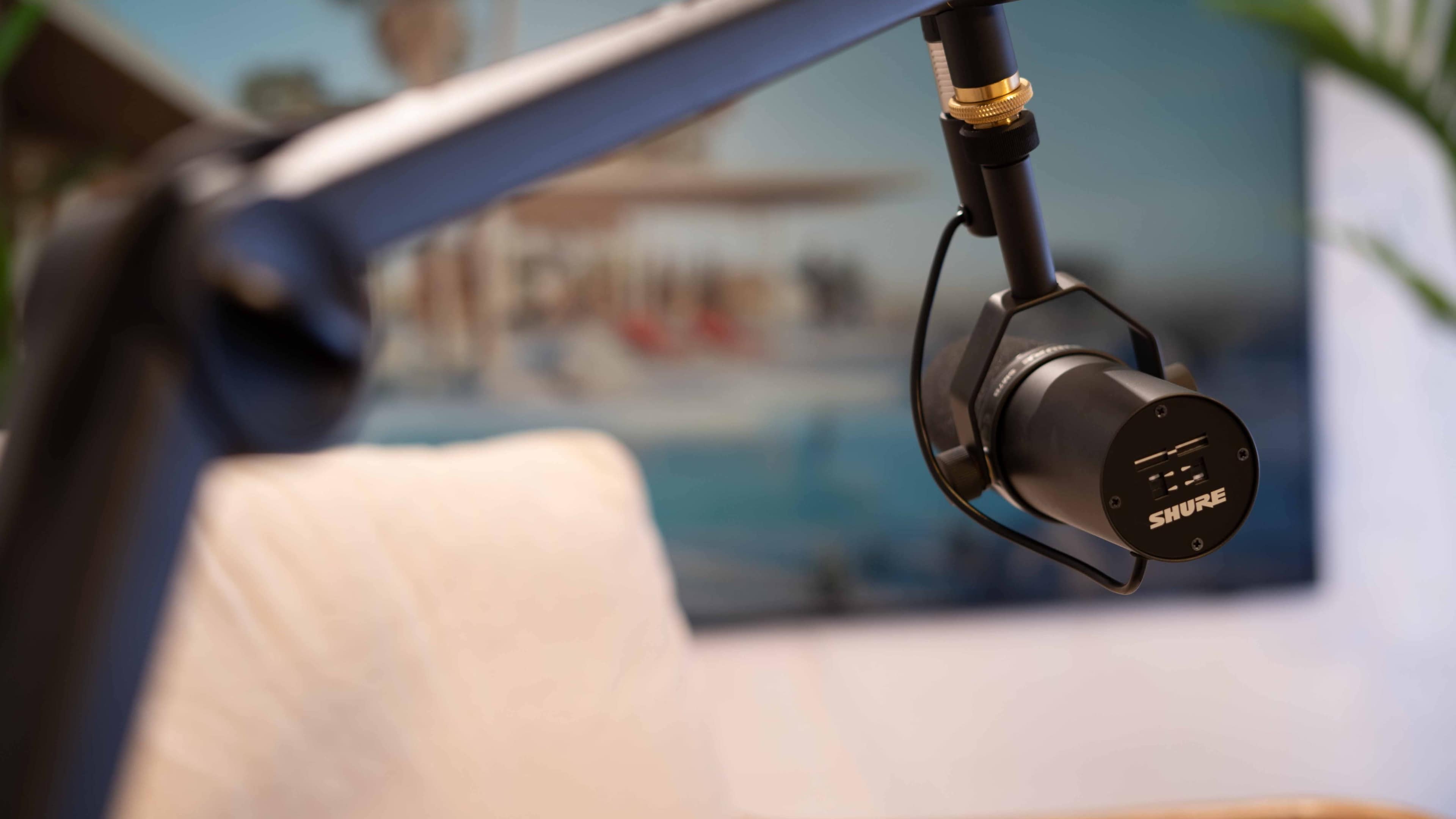A black microphone is mounted on an adjustable arm, positioned in front of a blurred background featuring a coastal scene.
