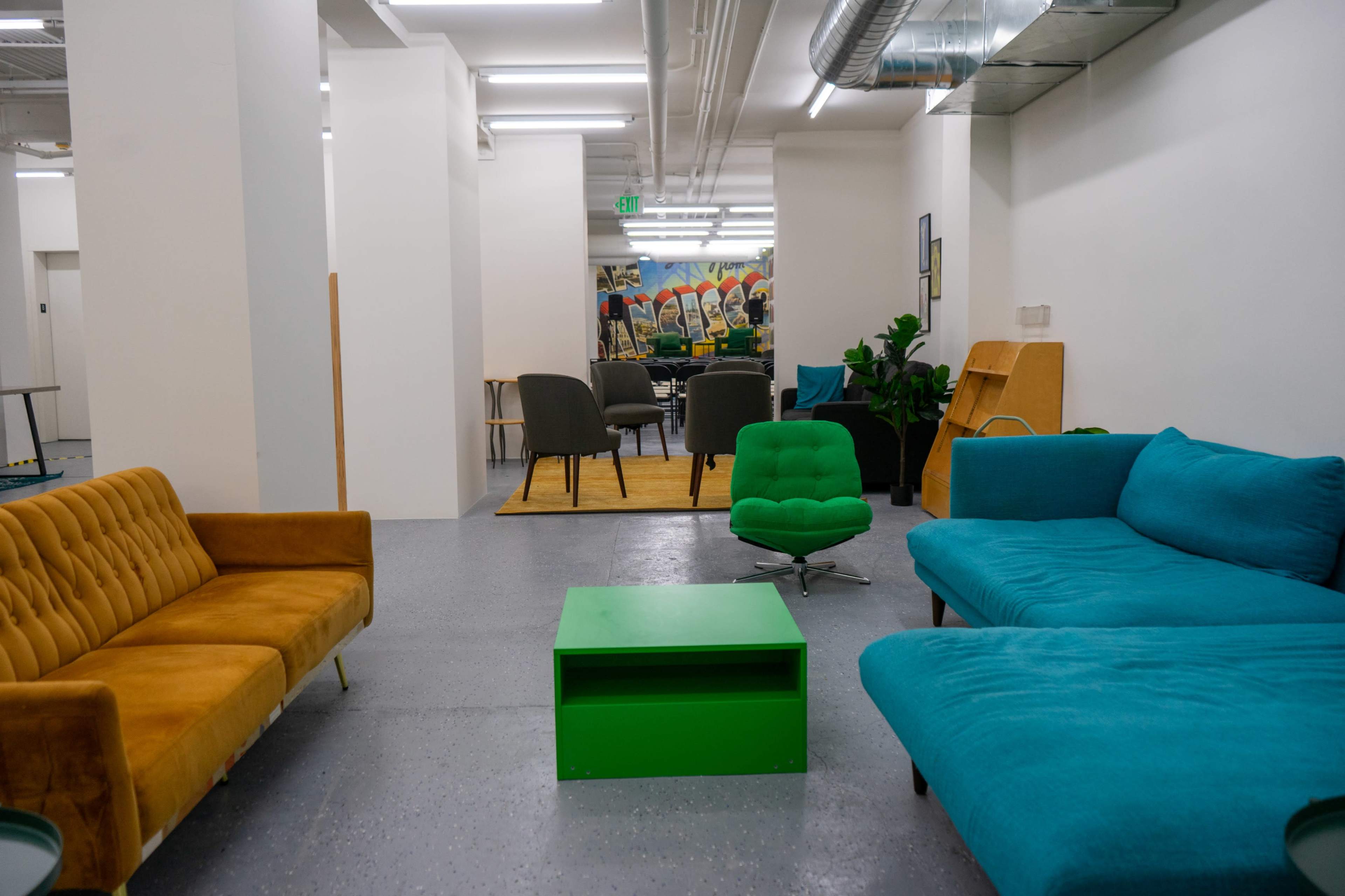 A modern, open lounge area features colorful seating, including an orange sofa and a teal sectional, along with a green coffee table and a backdrop of patterned murals.