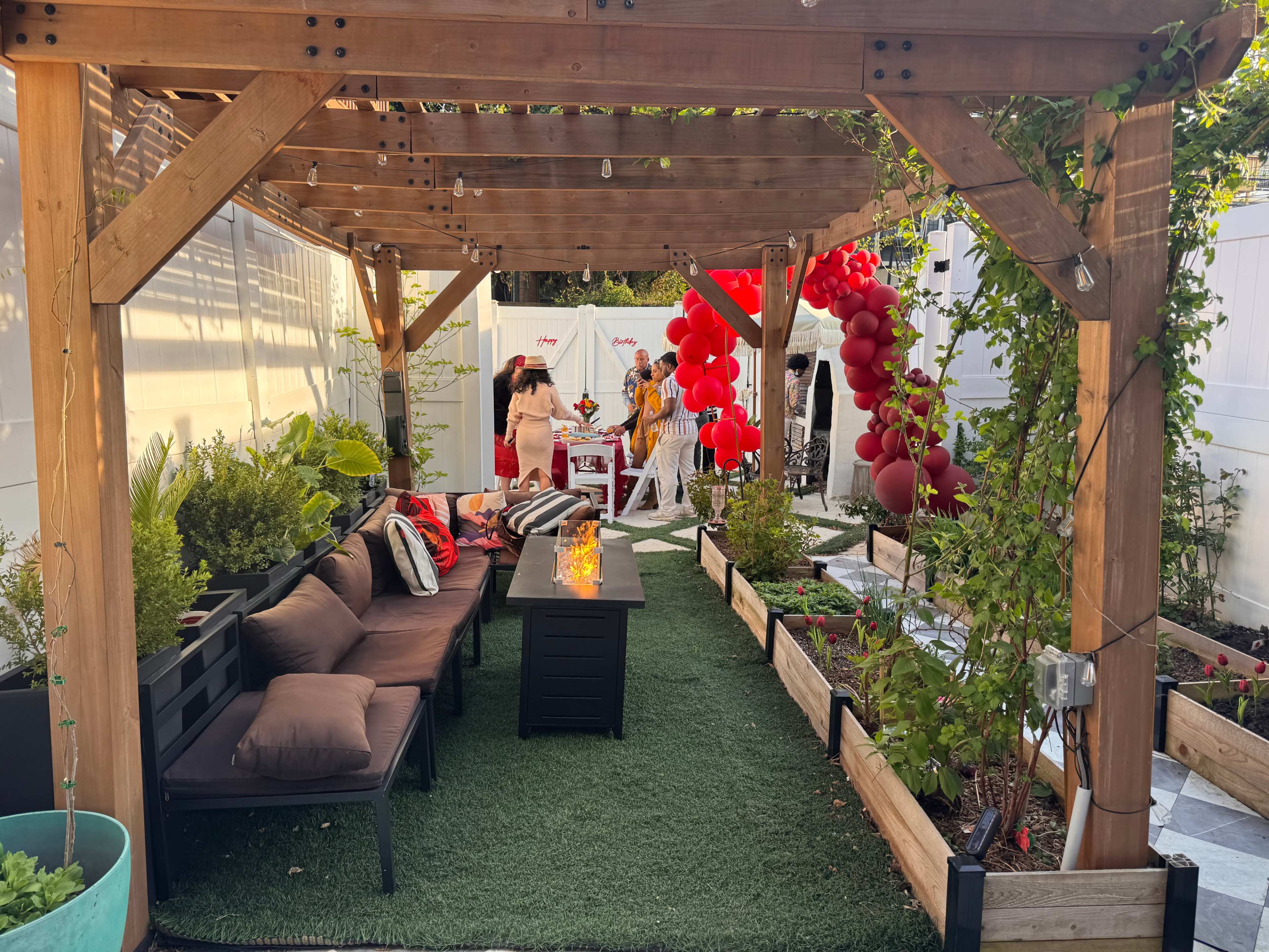 A shaded outdoor area features a wooden pergola with seating, decorative plants, and a vibrant balloon arch, set up for a gathering.