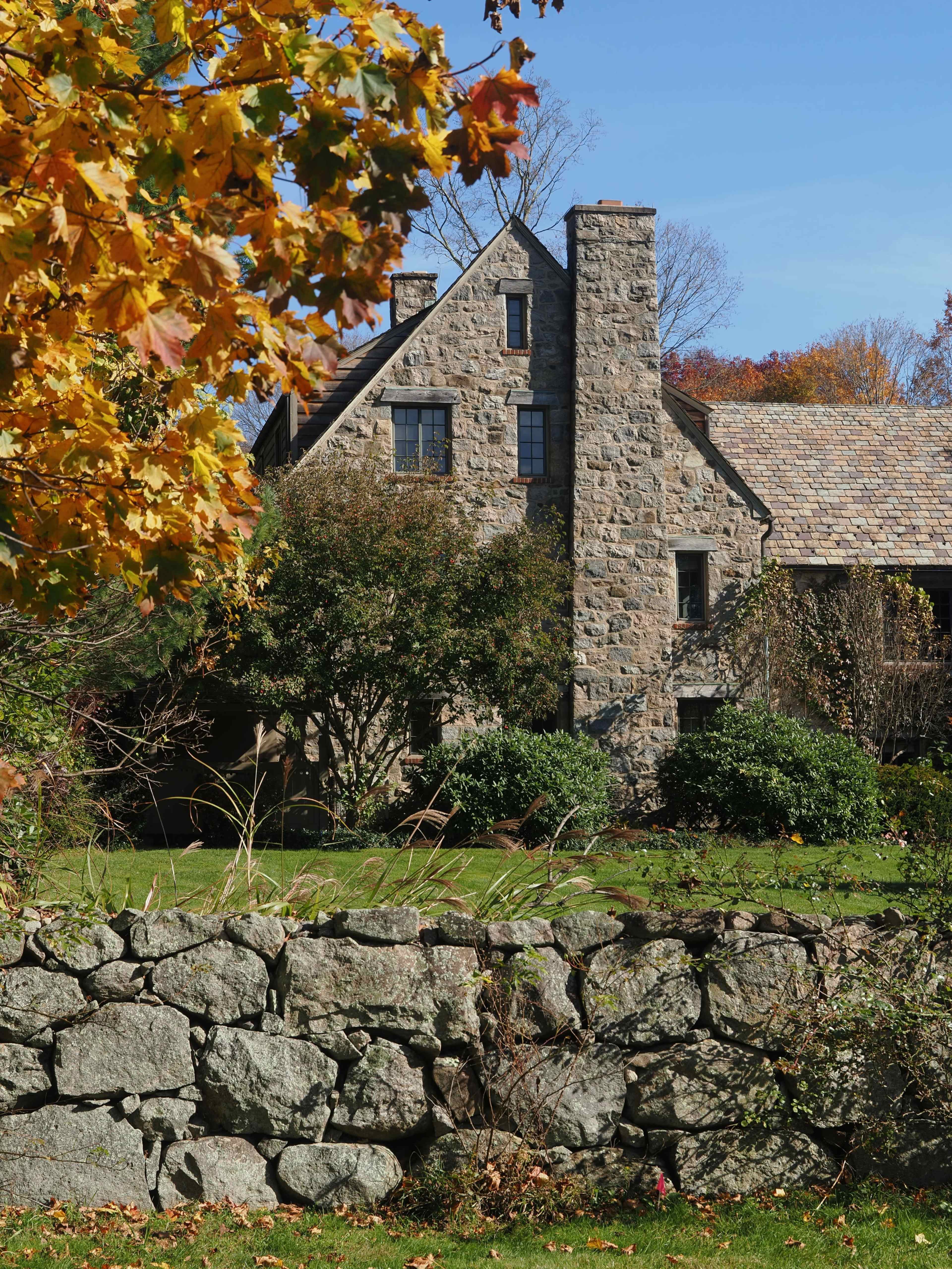 A stone house with a chimney is surrounded by greenery and a dry stone wall, under a clear blue sky with autumn foliage nearby.