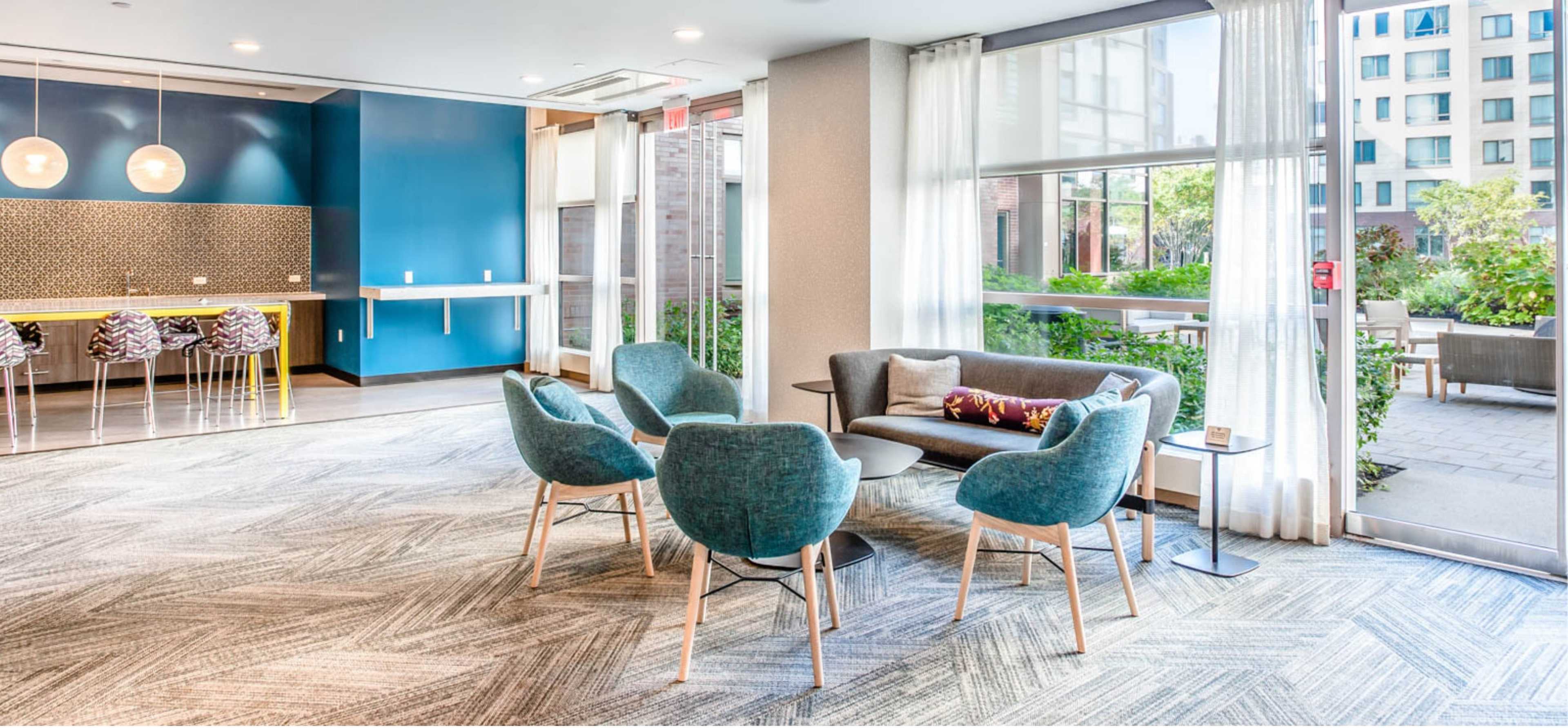 A modern lounge area with a seating arrangement of light blue chairs and a gray sofa, set against a backdrop of large windows and a colorful accent wall.