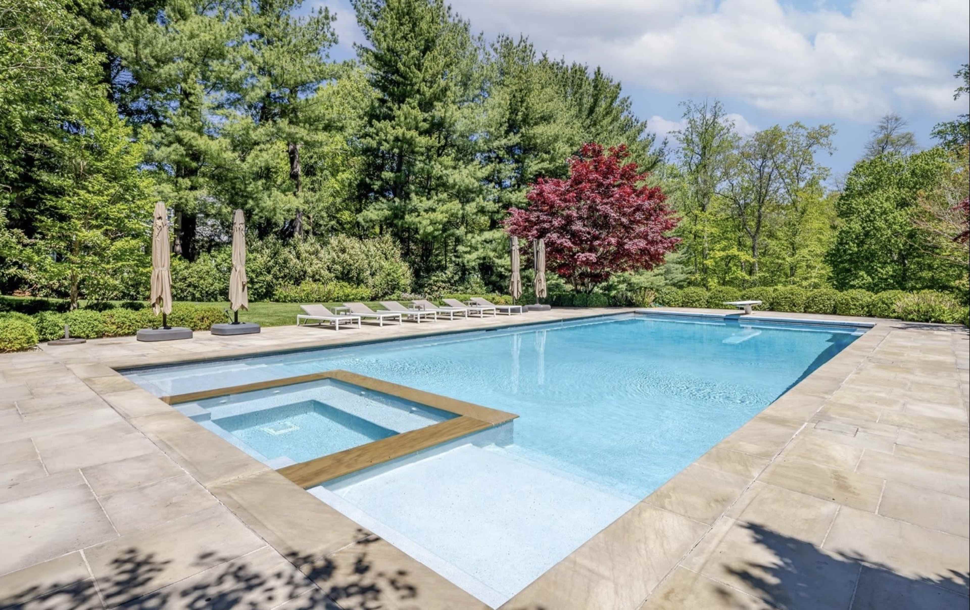 The image shows a rectangular swimming pool surrounded by stone tiles, with lounge chairs placed nearby and trees in the background.