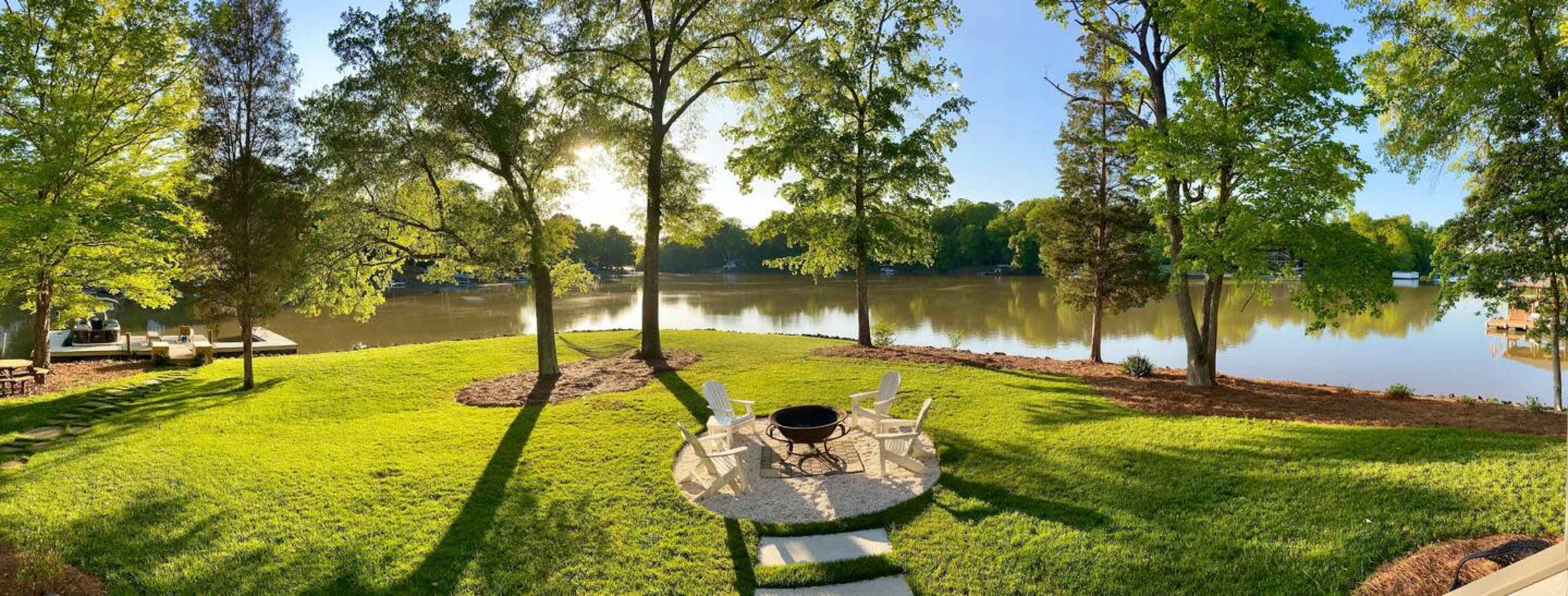 A landscaped yard by a calm lake, featuring a fire pit surrounded by chairs and bordered by trees.