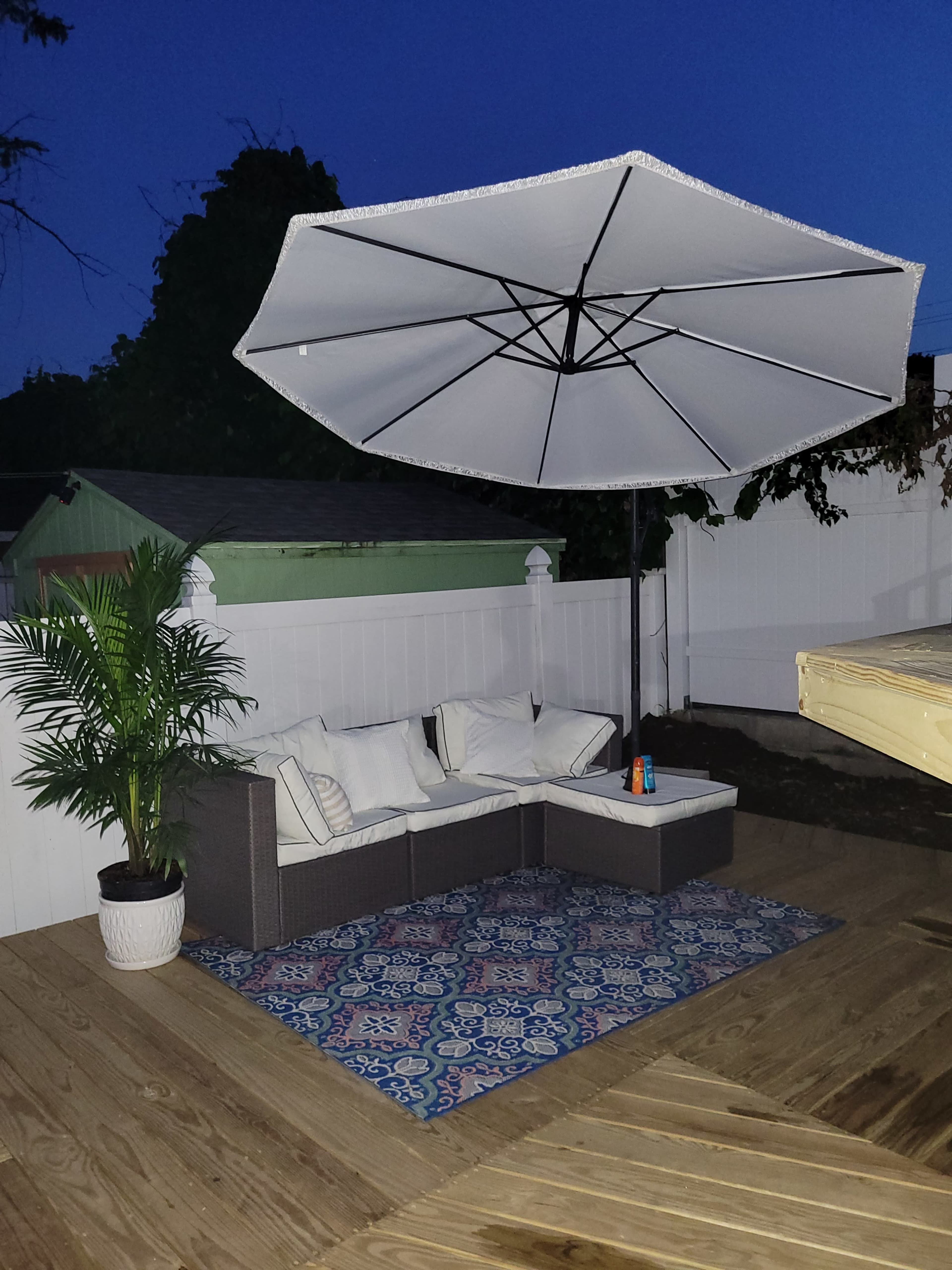 A covered seating area with a gray umbrella, two white cushions, and a blue decorative rug is set on a wooden deck surrounded by a white fence.