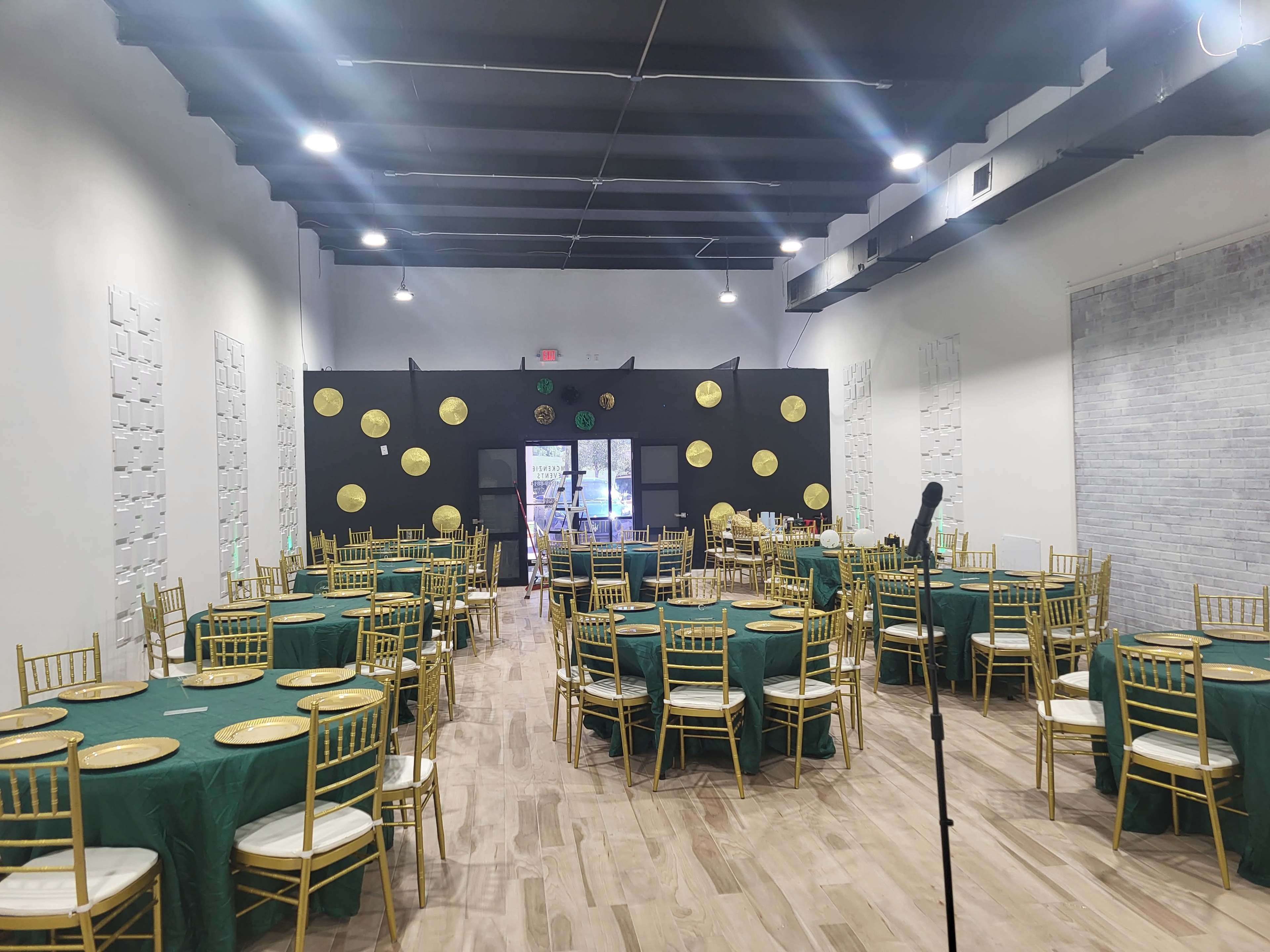 The image shows a large event space set up with round tables covered in green tablecloths and gold chairs arranged in a banquet-style layout.