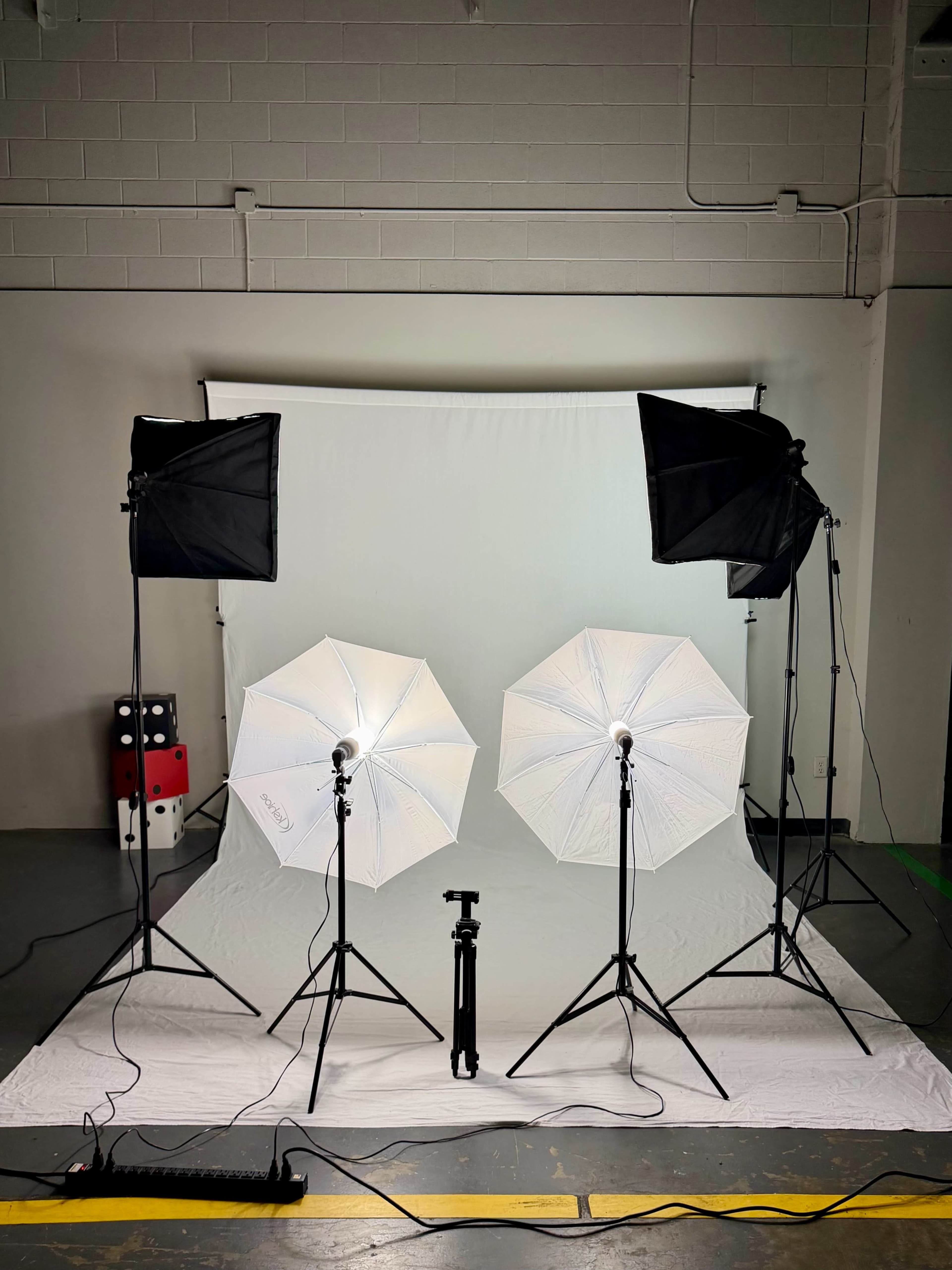 The image shows a photography studio setup with two softbox lights and two umbrella lights positioned in front of a white backdrop.