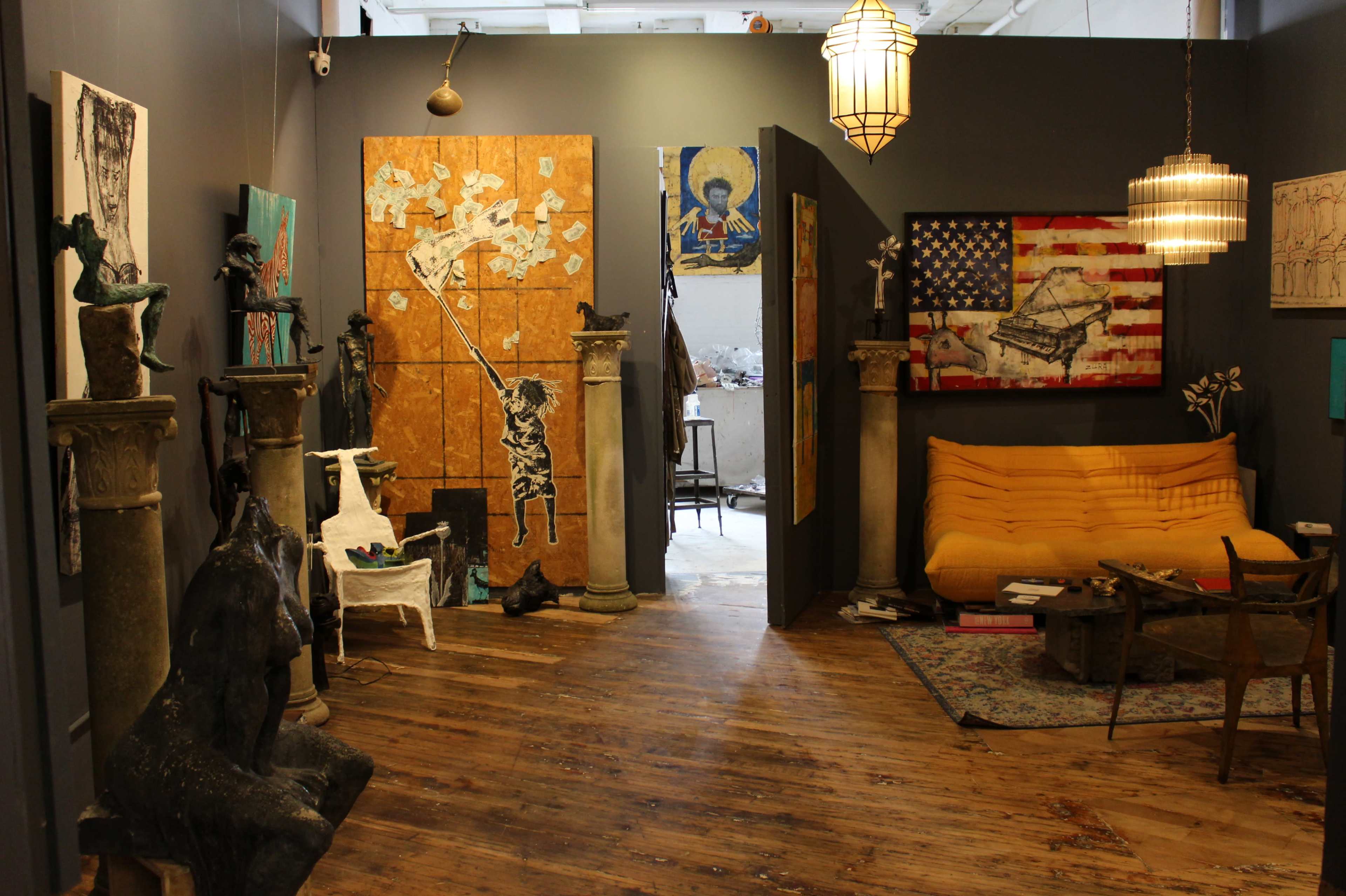 The image depicts an art gallery interior featuring various sculptures and paintings, with a yellow couch and wooden flooring.