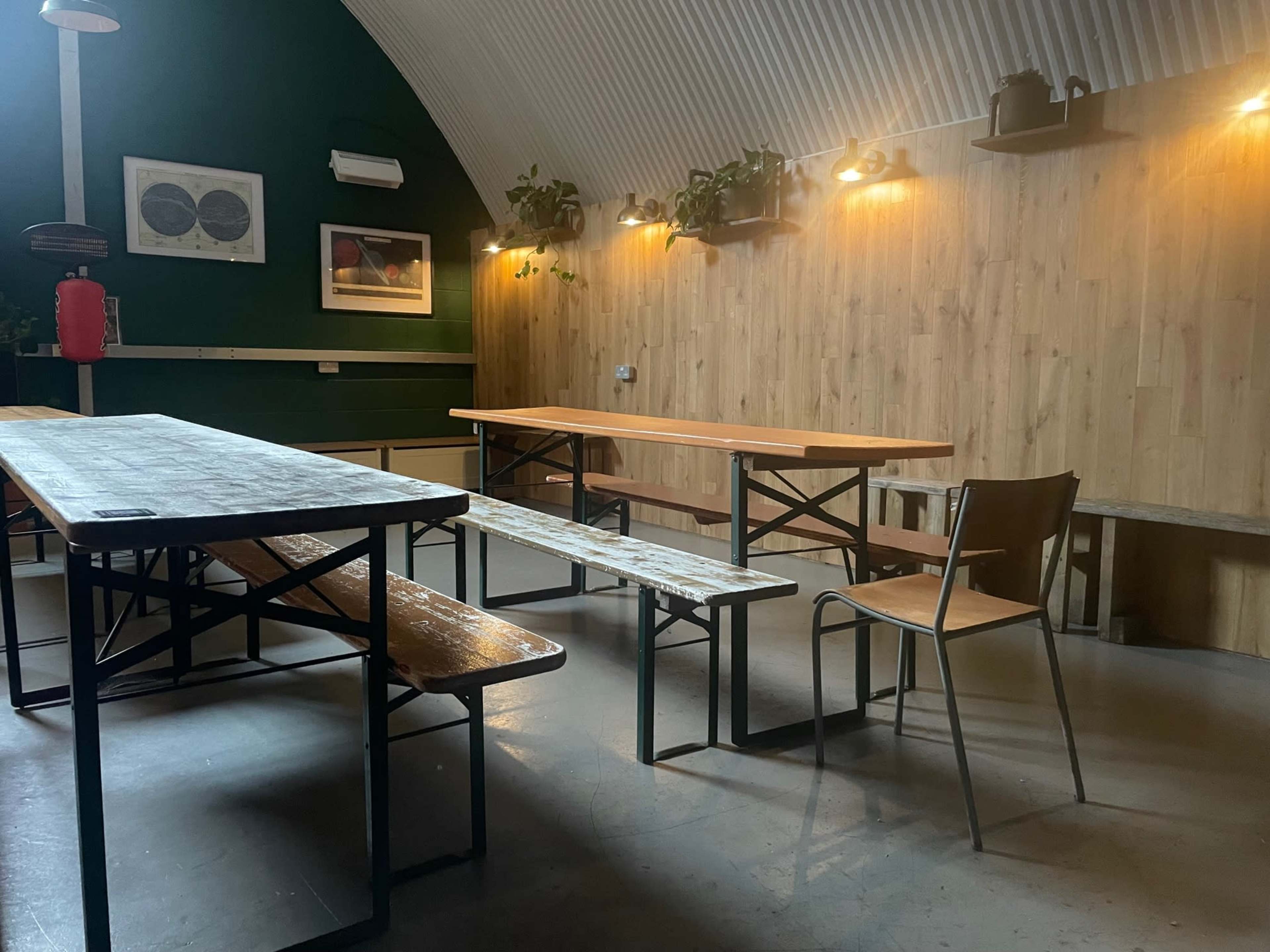 The image shows a minimalistic interior space with wooden tables and benches, a single chair, and potted plants against a wooden wall illuminated by warm lighting.