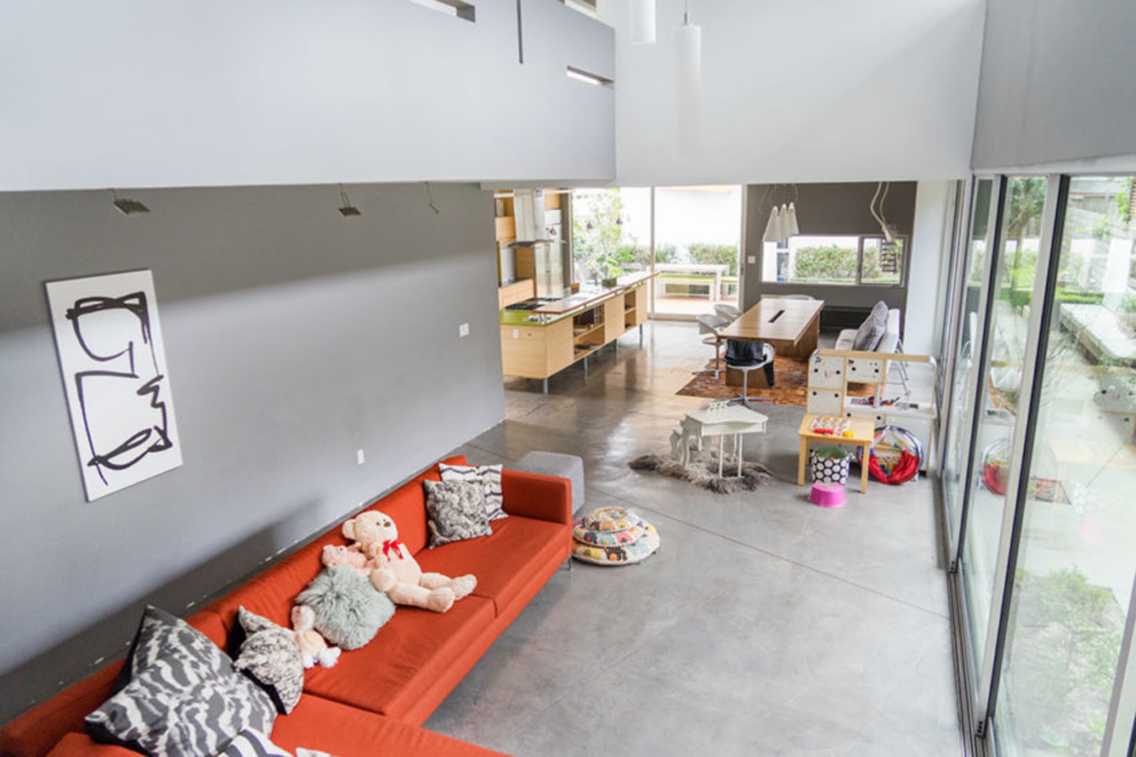 The image shows a modern, open-plan living area featuring a red sofa, a dining table, and a kitchen with an island, all surrounded by large windows.