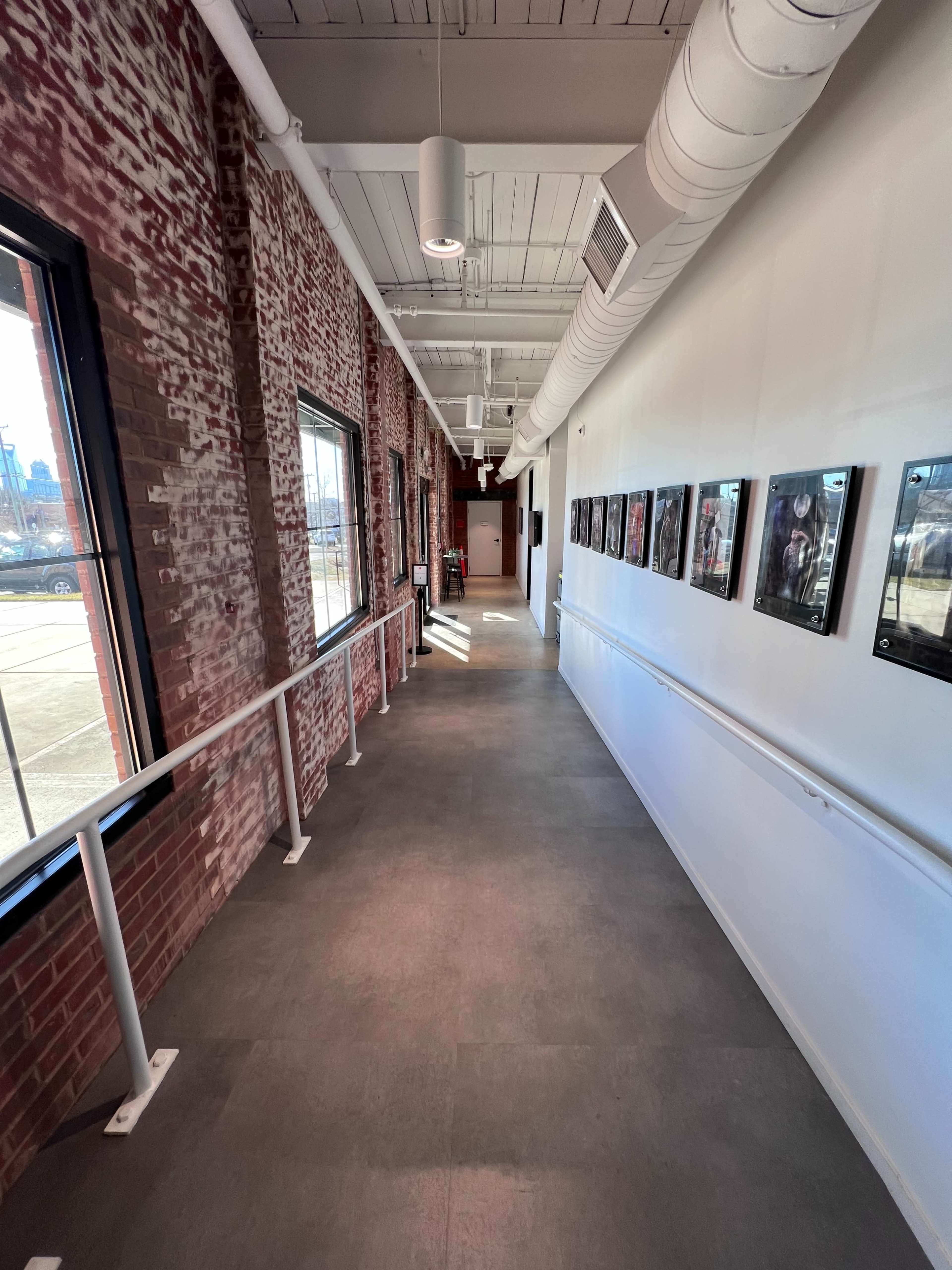 The image shows a long hallway with brick walls, framed photos on one side, and a smooth floor leading to a door at the end.
