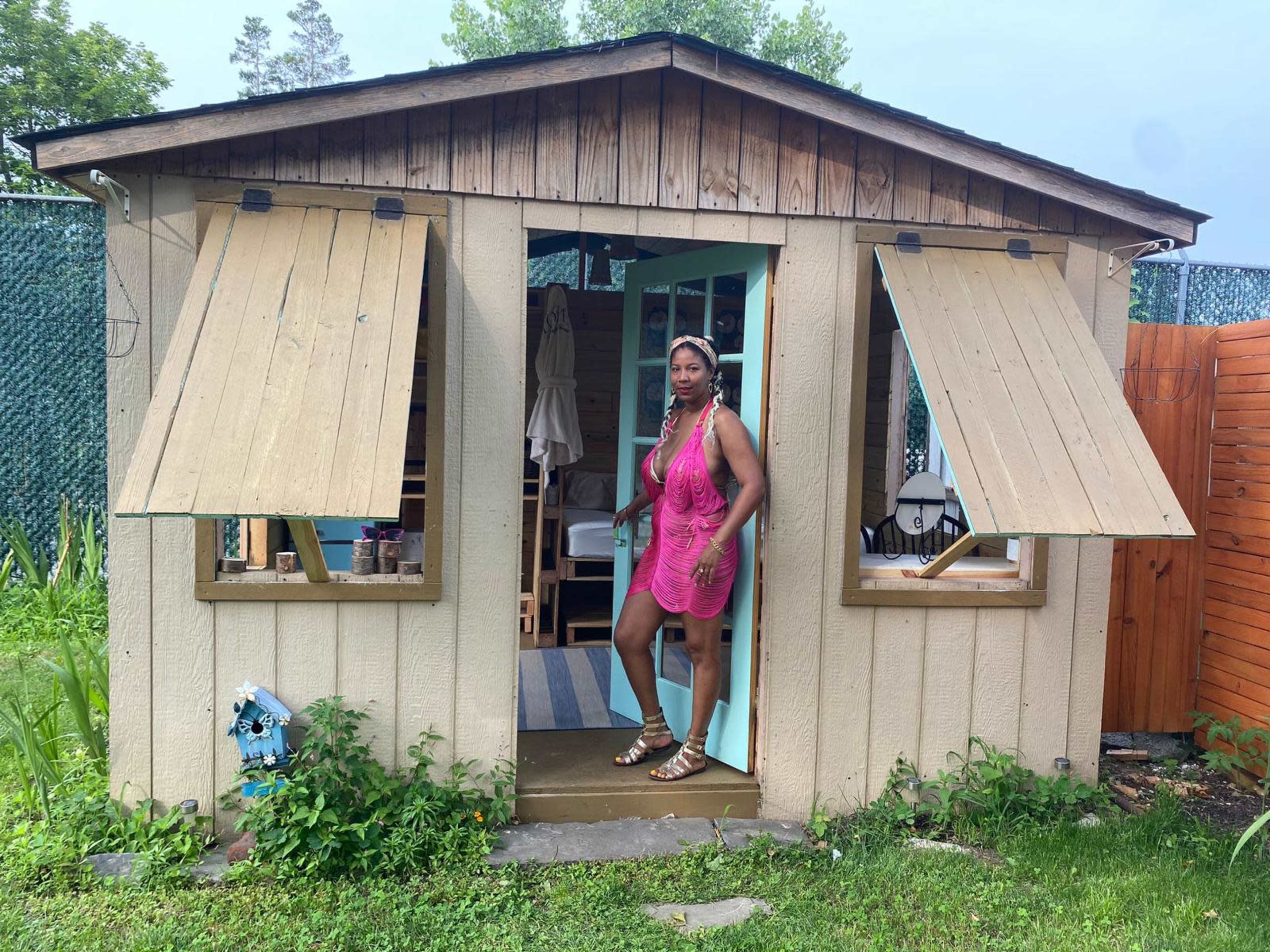 A woman stands in the doorway of a small, wooden shed with open shutters, surrounded by greenery.