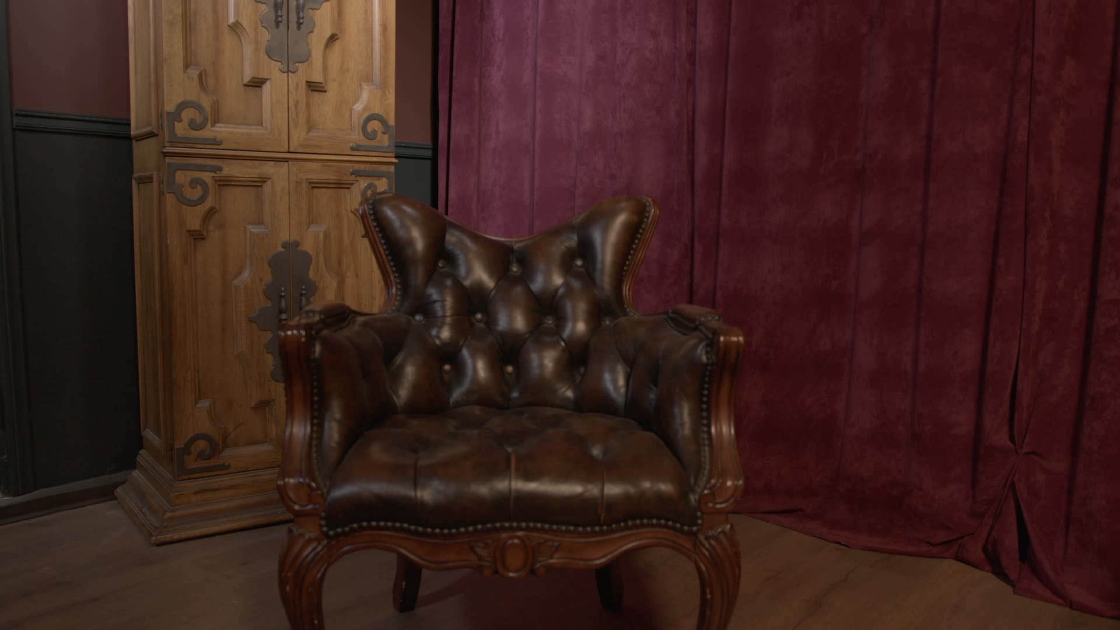 The image shows a brown leather armchair with a tufted design, positioned in front of a purple fabric backdrop and a wooden cabinet.