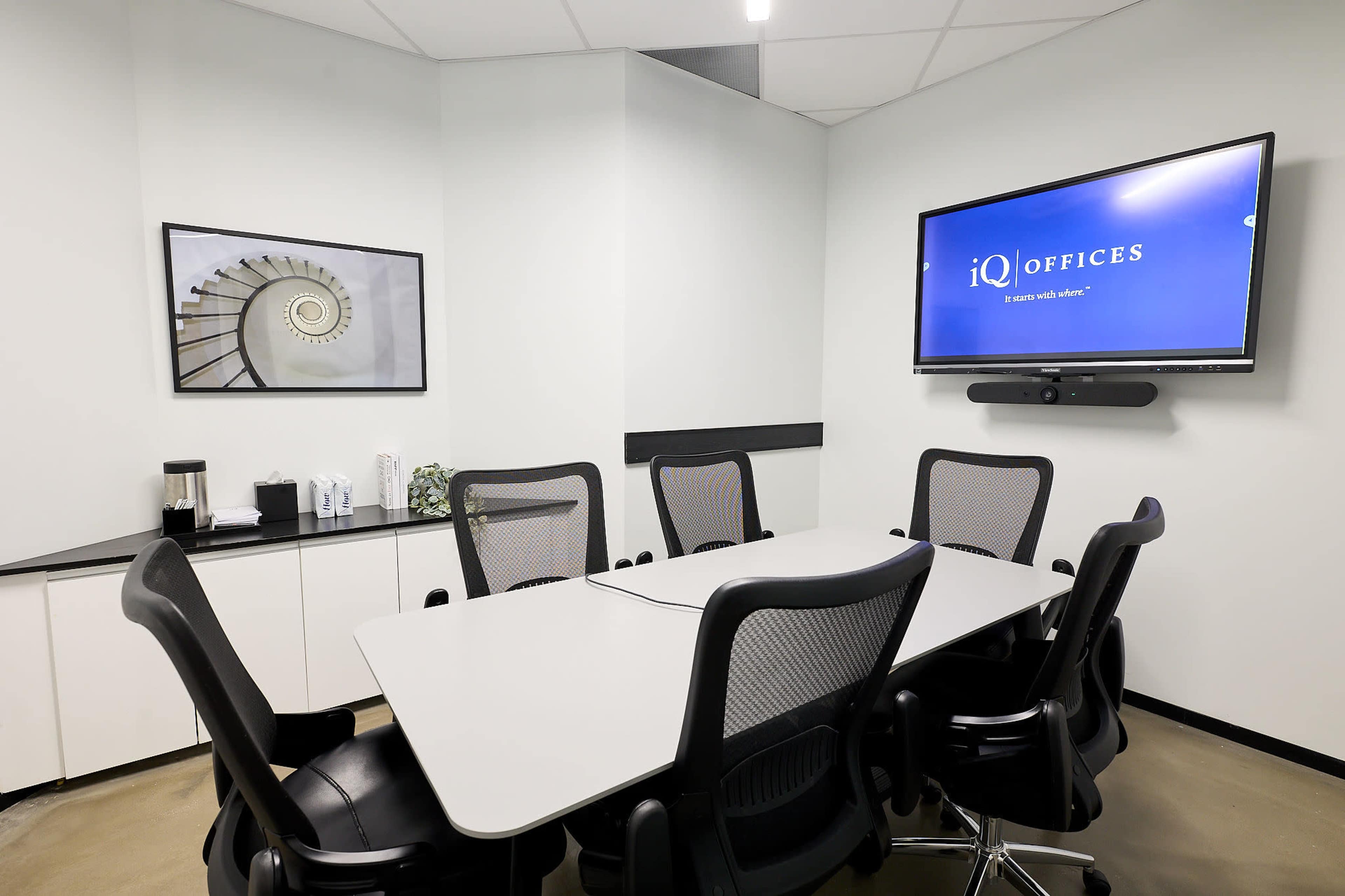 The image shows a modern conference room equipped with a large table, several ergonomic chairs, and a wall-mounted television displaying the iQ Offices logo.