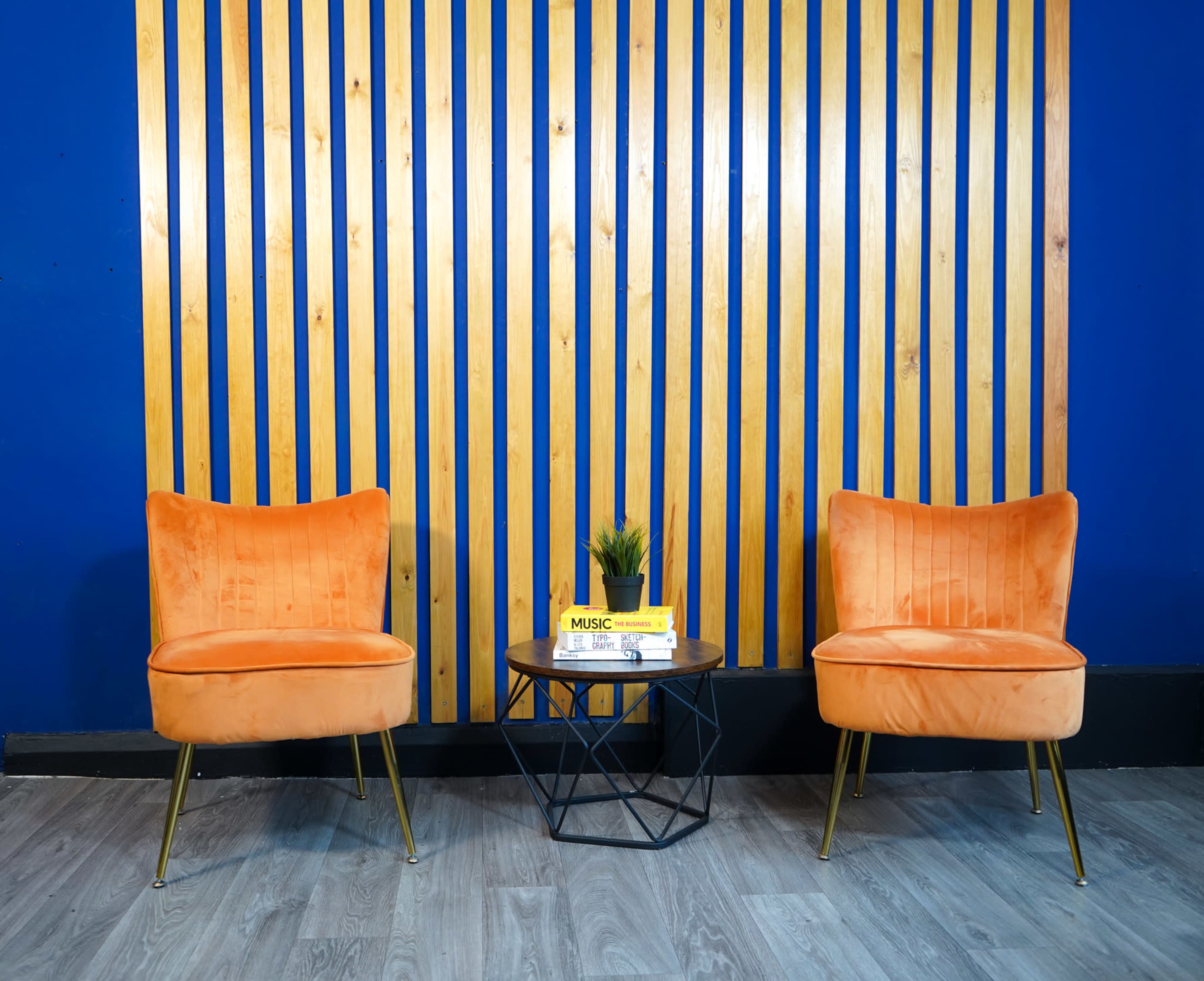 Two orange chairs are positioned on either side of a round table with a small plant, set against a blue wall featuring vertical wooden slats.