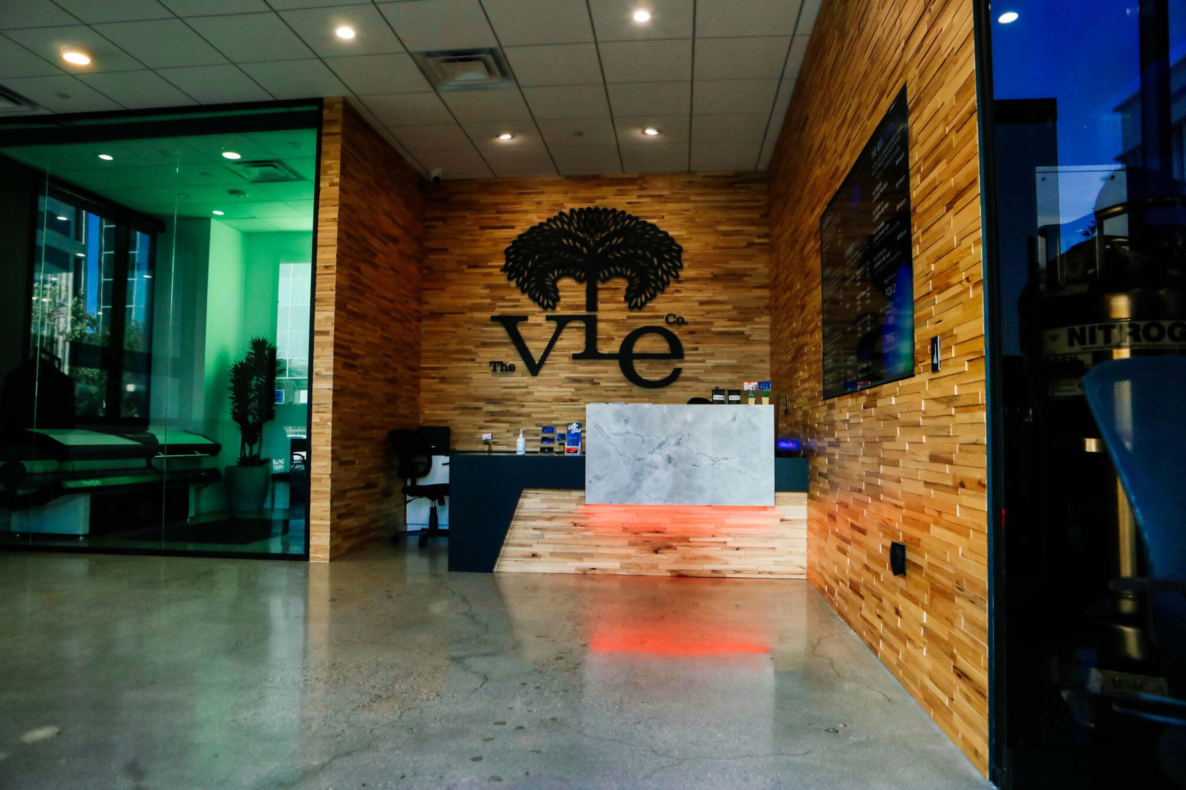 The image shows a modern reception area featuring a wooden wall with a logo, a marble counter with ambient lighting, and large glass windows.