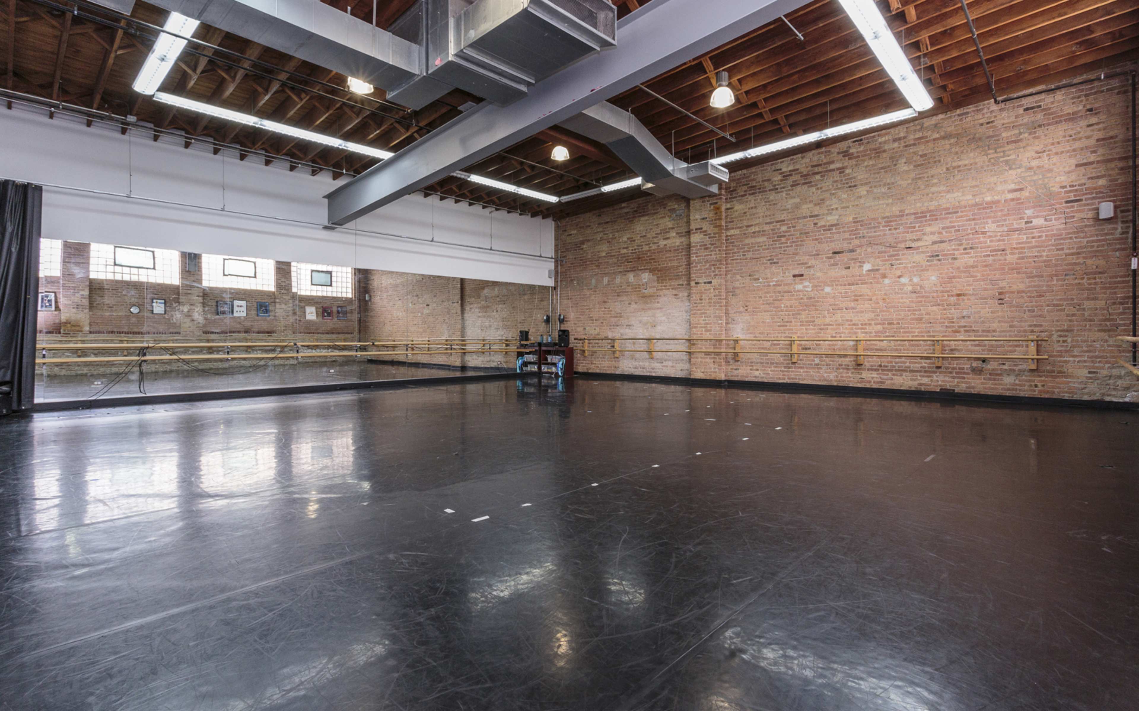 A spacious dance studio with a wooden ceiling, exposed brick walls, and a large mirror along one side.