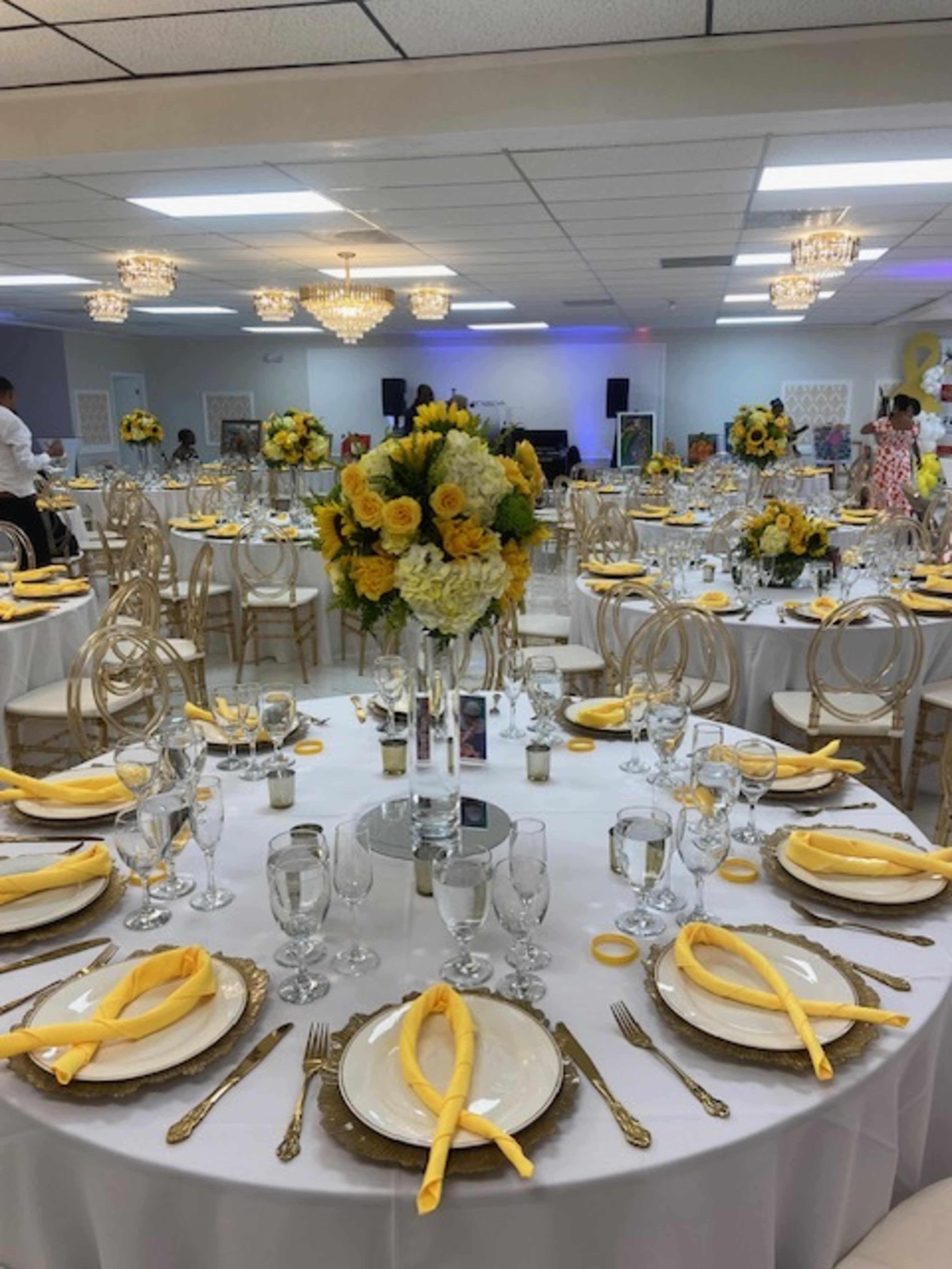 The image shows a banquet hall set up for an event, featuring tables elegantly arranged with white tablecloths, gold accents, and floral centerpieces of yellow flowers.