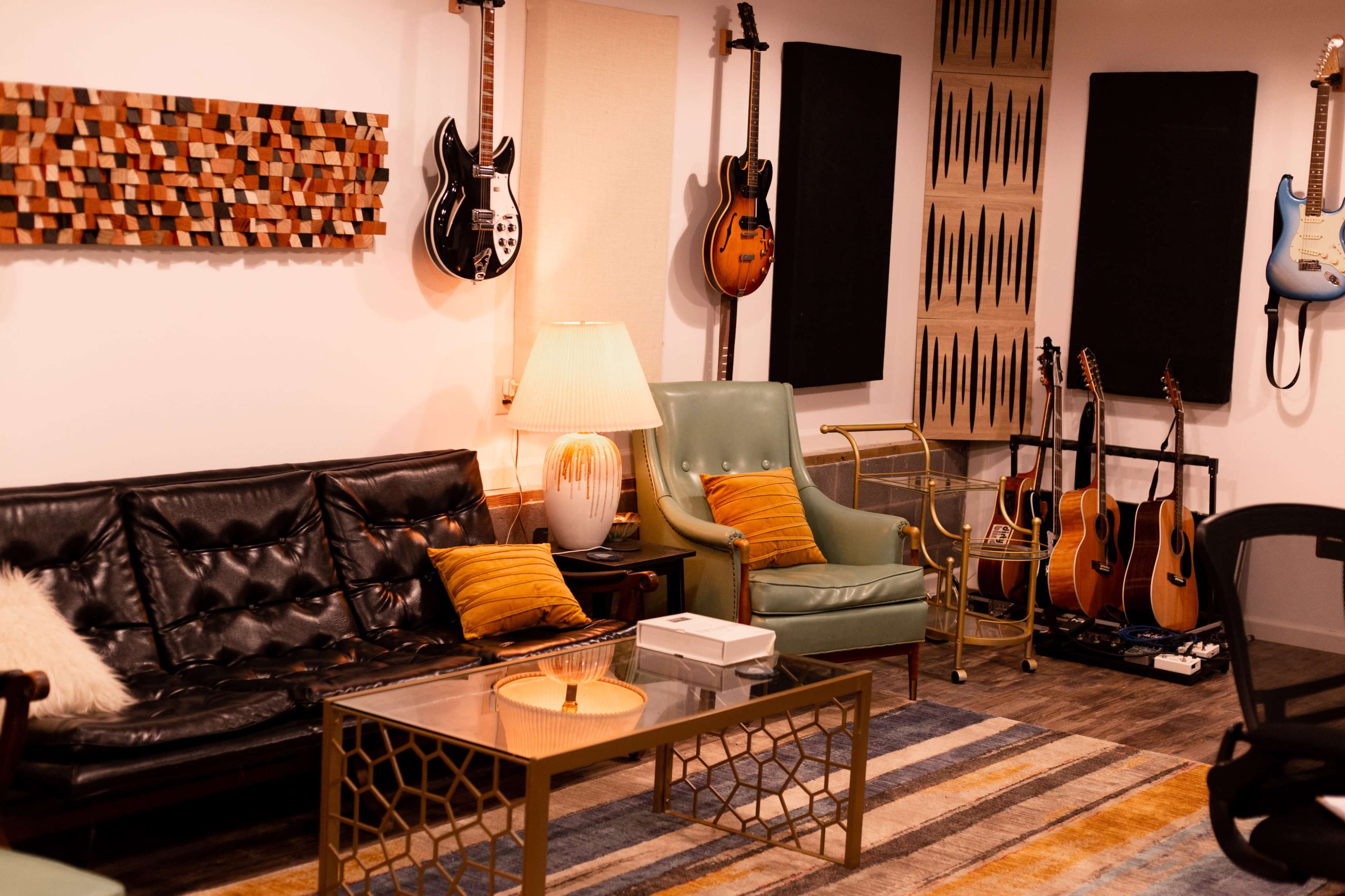 A cozy music studio featuring a black leather sofa, a green armchair, a coffee table, and various guitars displayed on the walls.