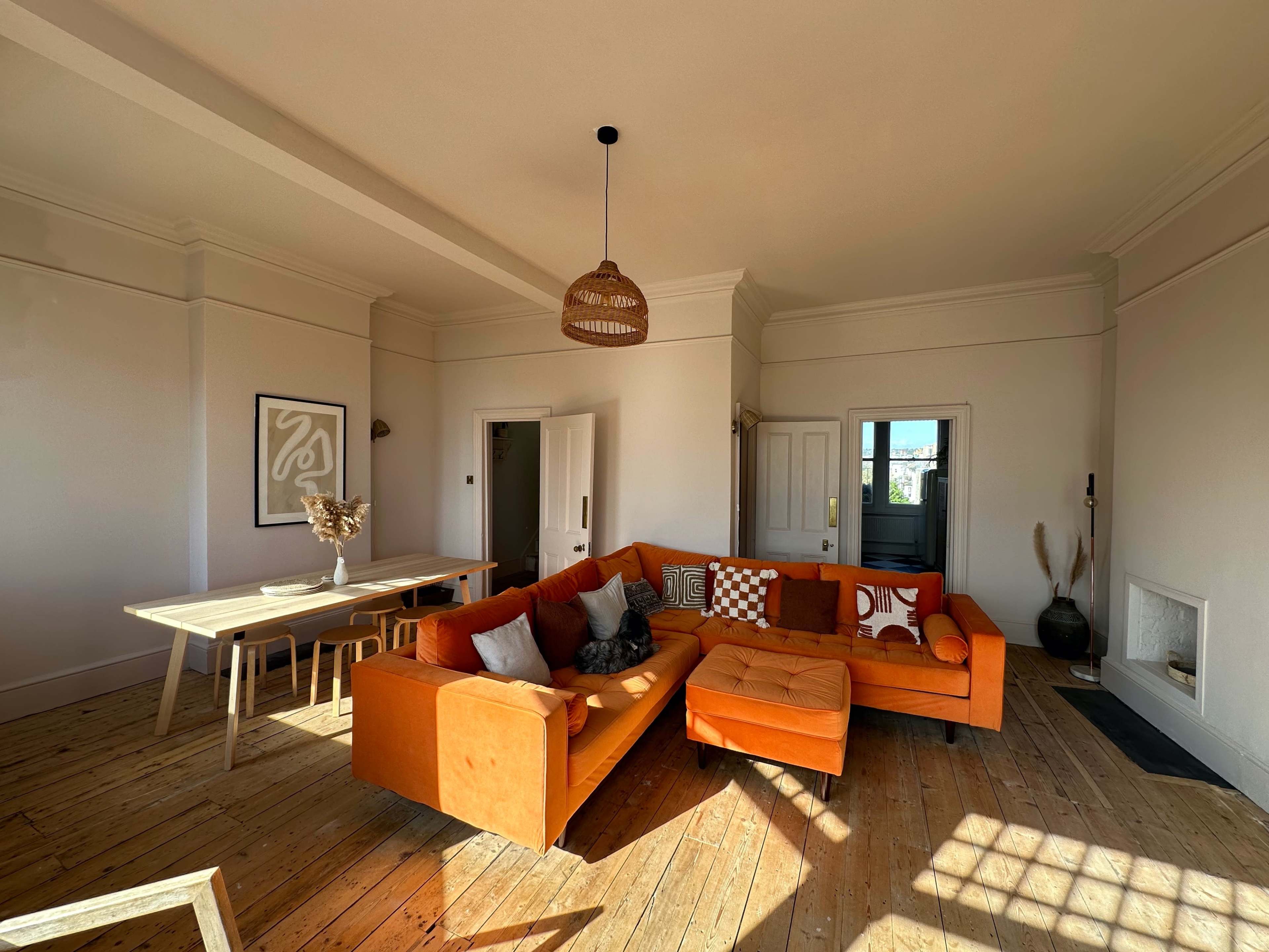 A living room with an orange sectional sofa, a wooden dining table, and various decor elements against a light-colored wall.