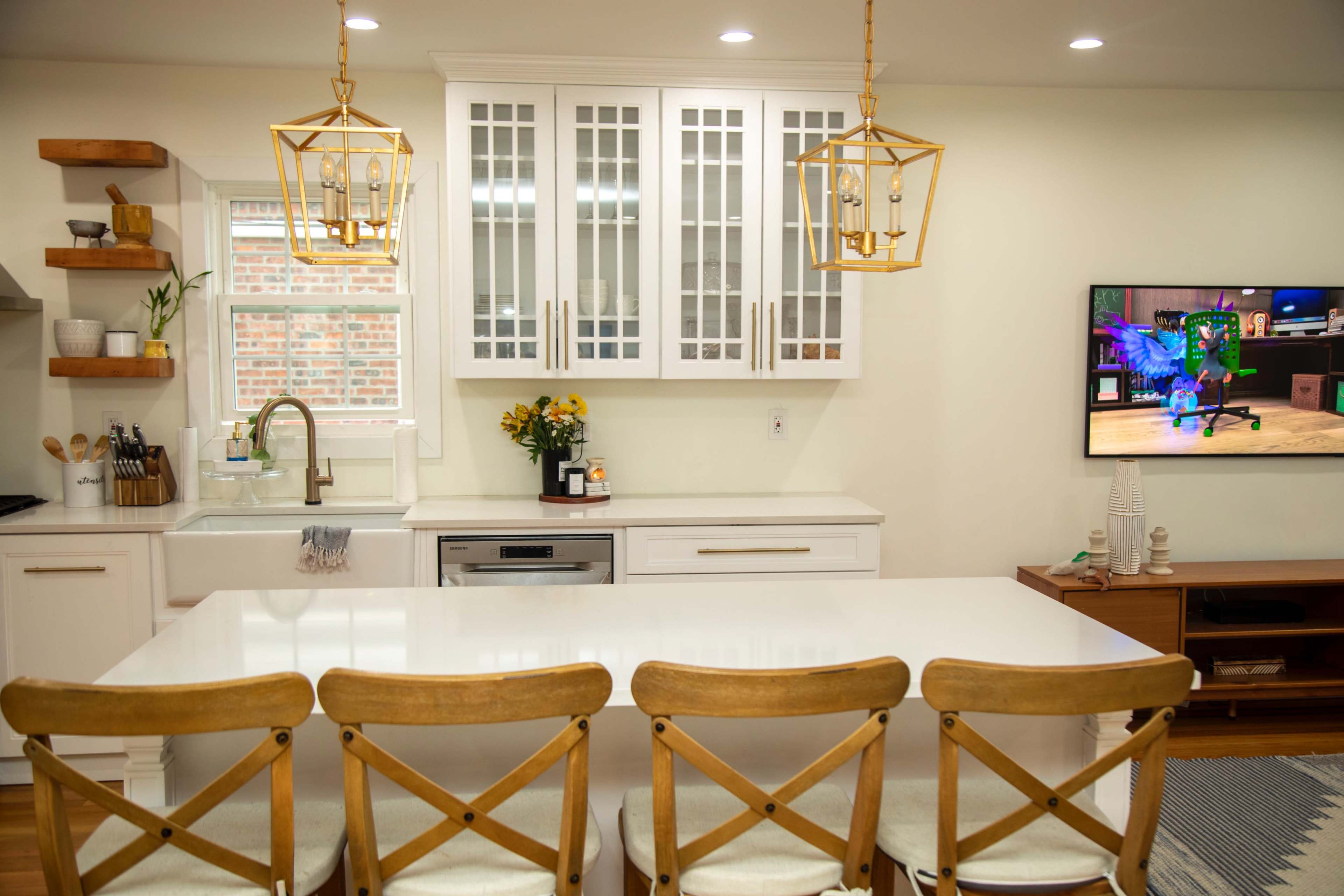 A modern kitchen features a central island with wooden chairs, white cabinetry with glass doors, and a television mounted on the wall displaying a colorful image.