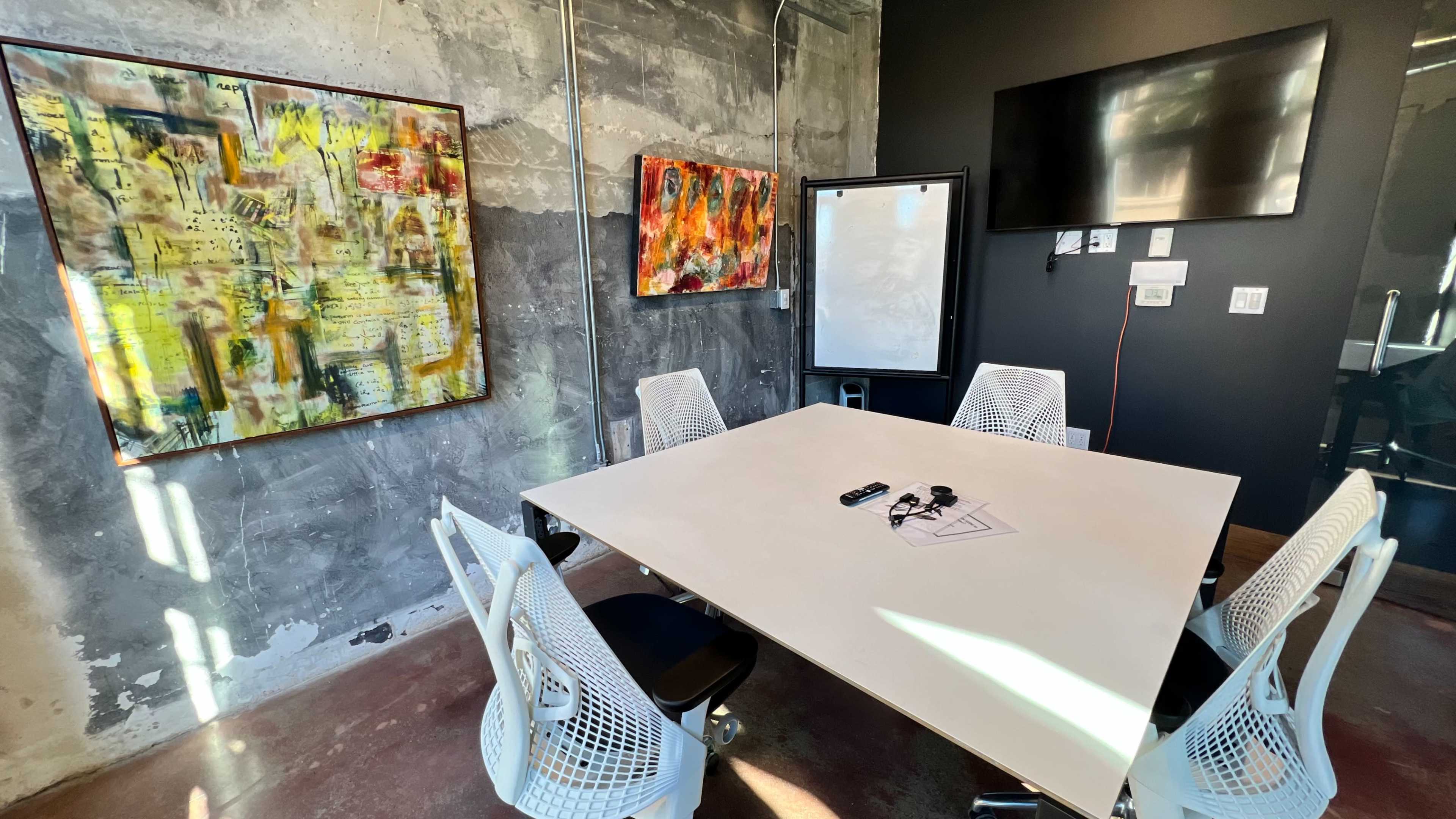 A modern meeting room features a white table surrounded by four white chairs, with abstract artwork on the walls and a wall-mounted screen.