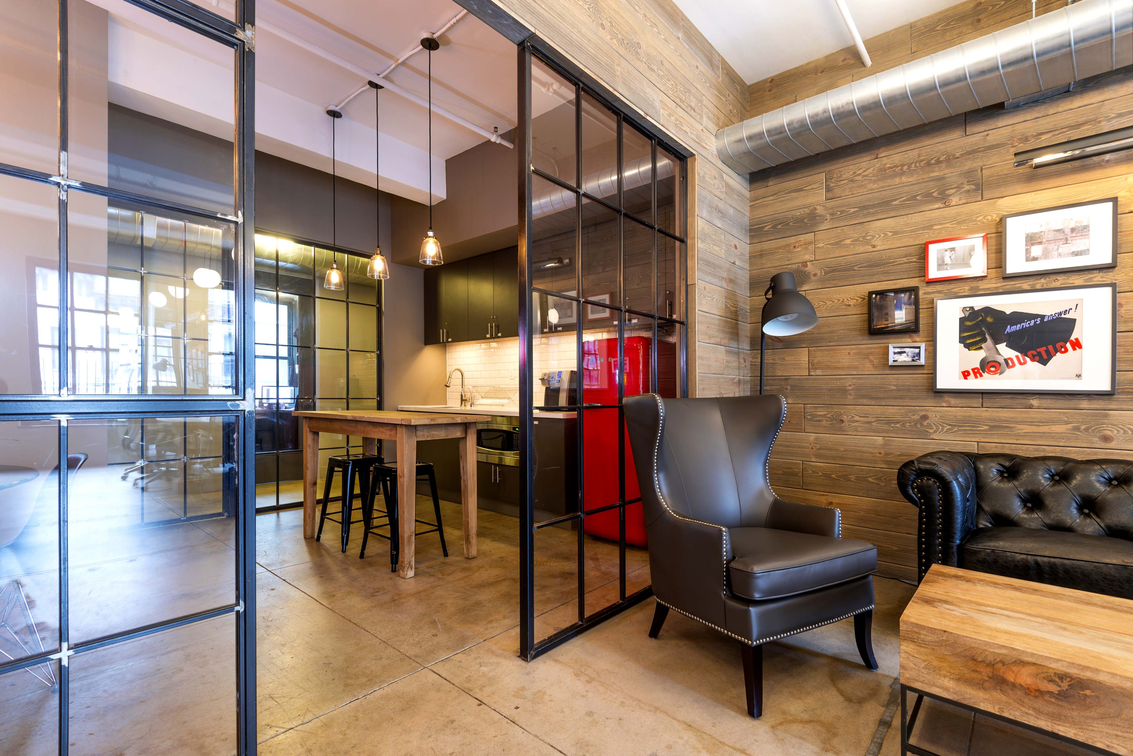 The image shows a stylish office space with a glass partition, featuring a sitting area with a leather chair and a wooden table, along with a kitchenette and a red refrigerator in the background.