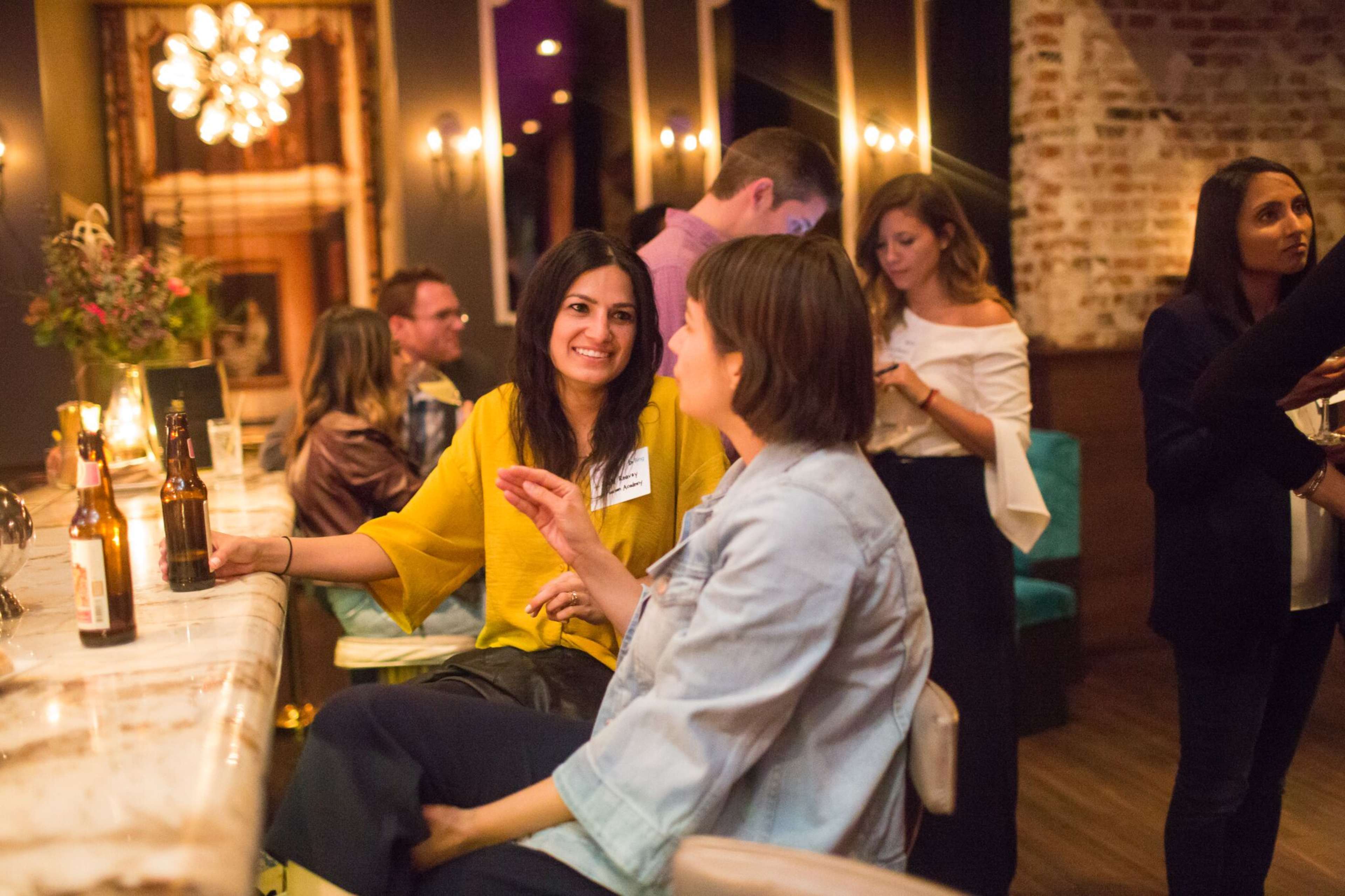 Two women sit at a bar, engaged in conversation while others socialize in the background.
