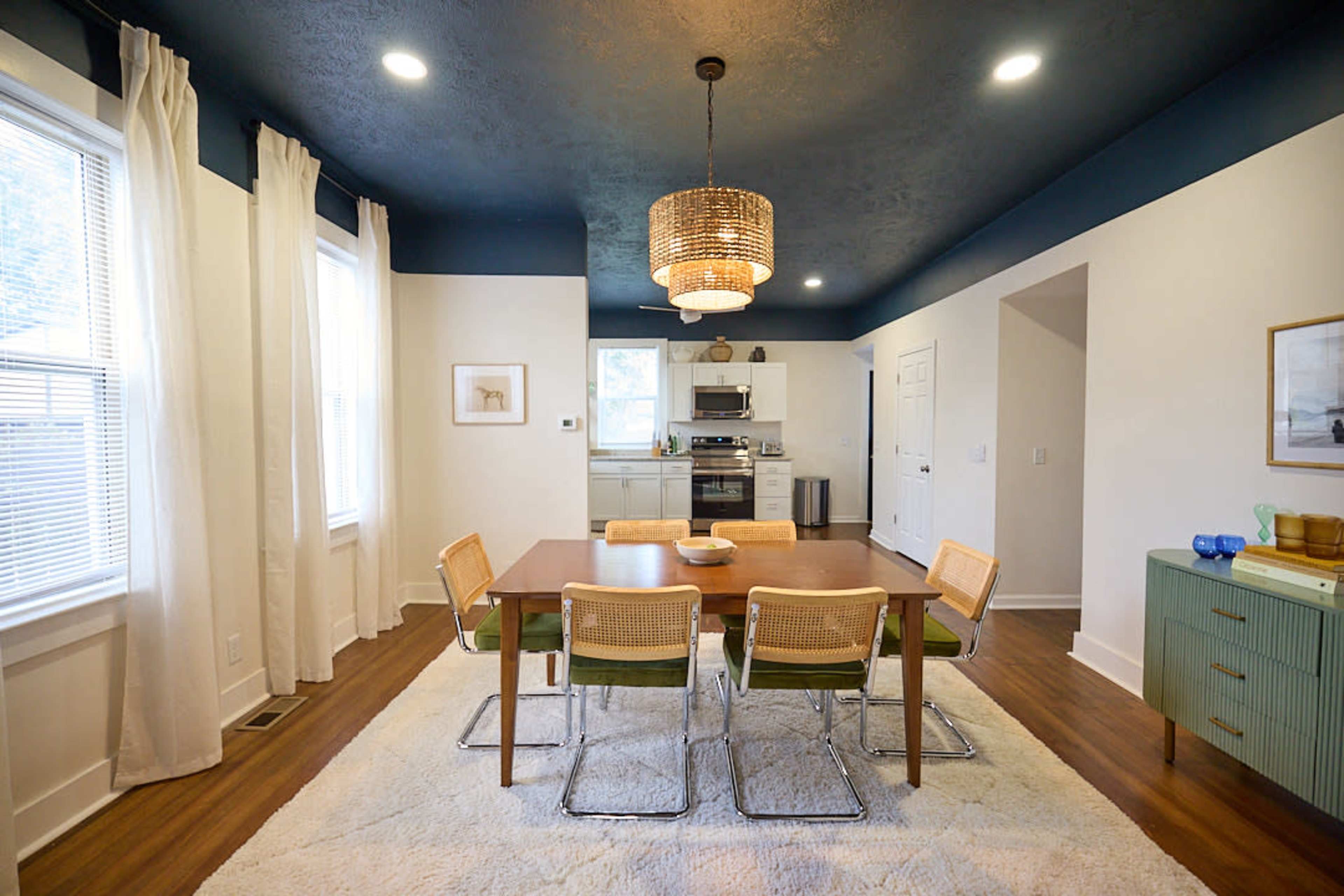 The image shows a modern dining area with a wooden table surrounded by four chairs, a light fixture hanging from the ceiling, and a kitchen visible in the background.