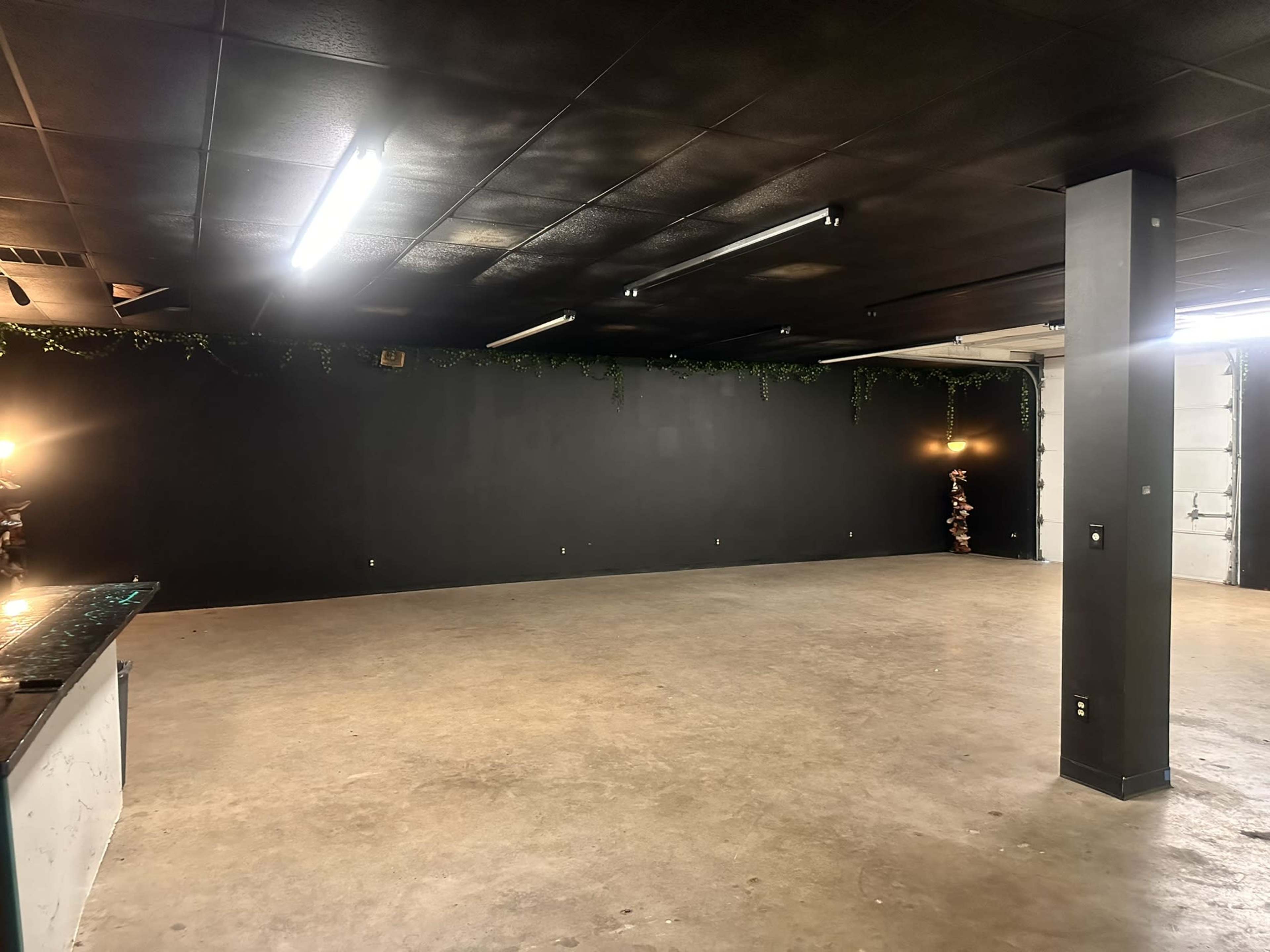 The image shows a large, empty room with a black ceiling and walls, featuring concrete flooring and minimal lighting from fixtures along the ceiling.