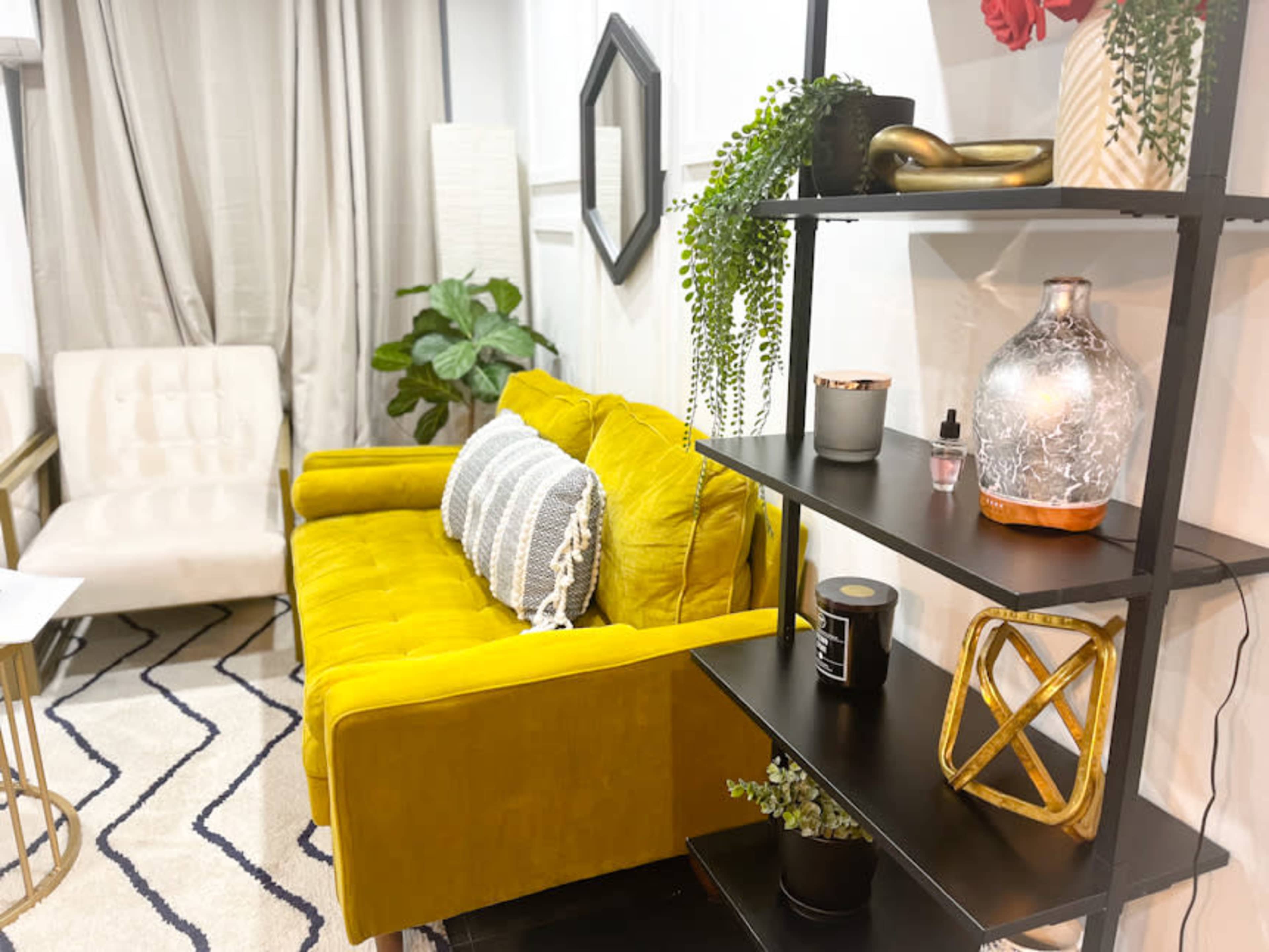 A bright yellow couch positioned near a black shelving unit filled with decor items, with a potted plant and a styled chair in the background.