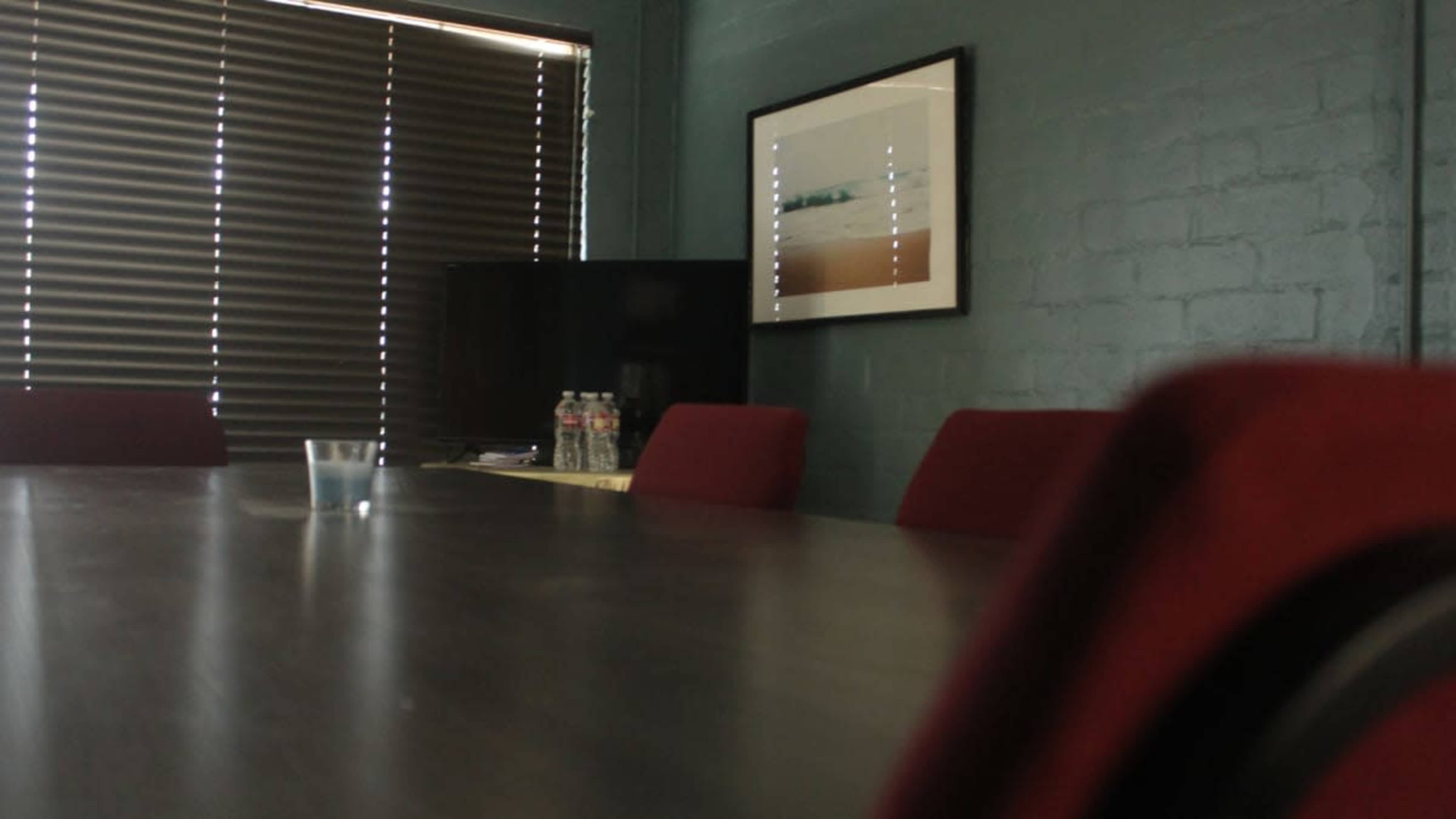 A conference room features a large table with red chairs, a blurred view of a television, and an art piece hanging on the wall, all illuminated by natural light from partially closed blinds.