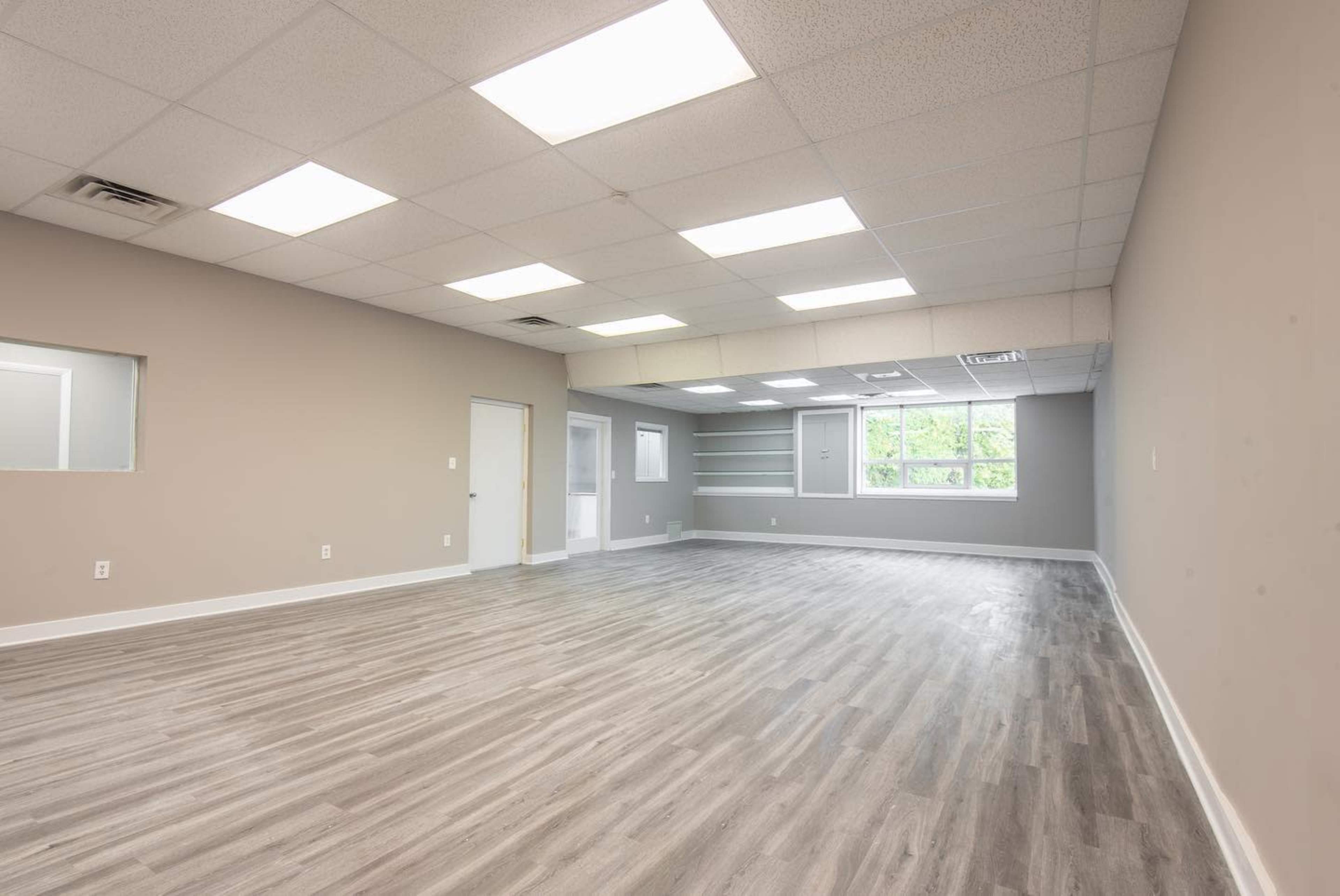 The image shows a large, empty room with light-colored walls and a wooden floor, featuring fluorescent ceiling lights and windows.