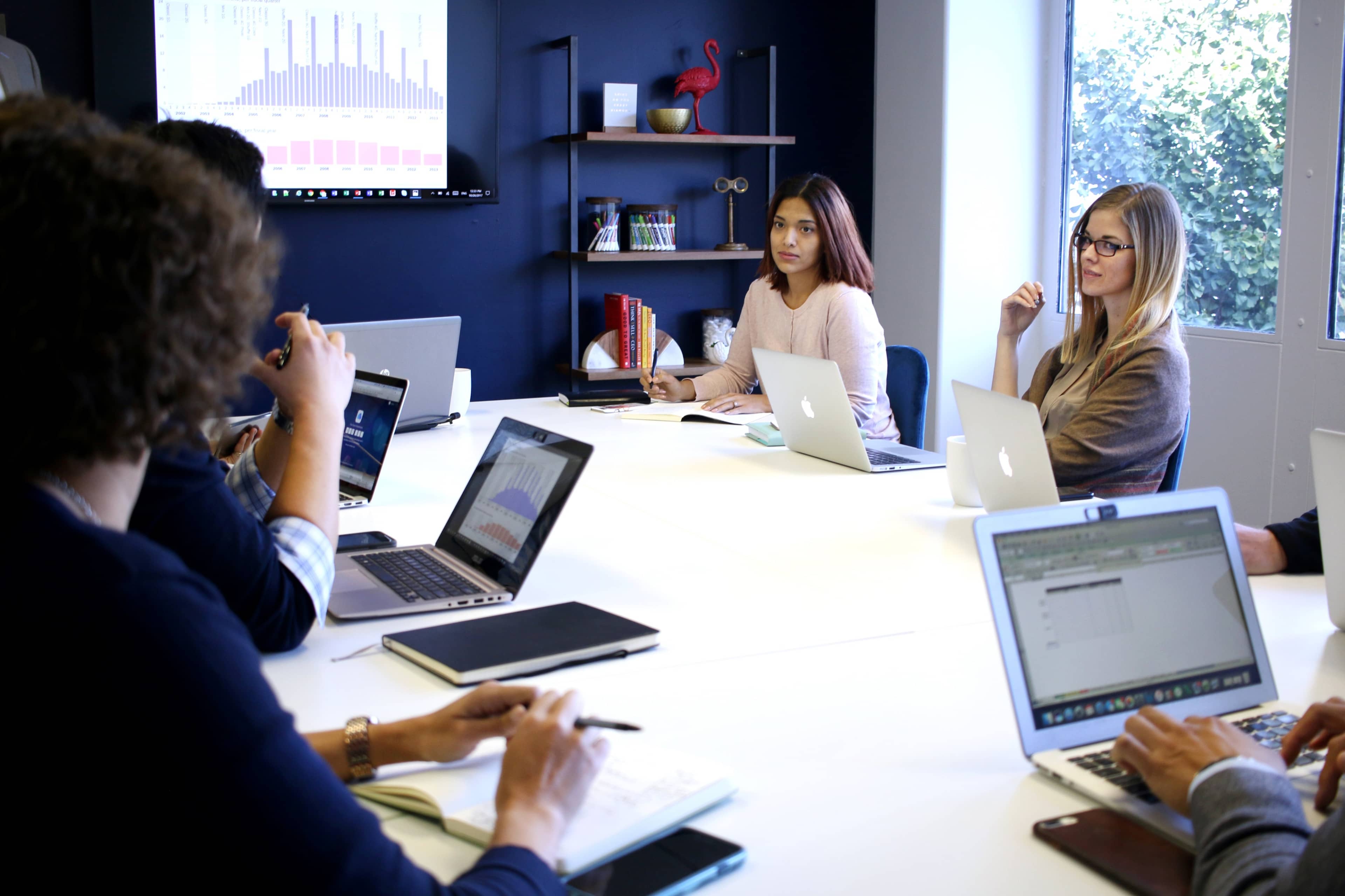 A group of people is seated around a conference table in a modern meeting room, engaged in discussion while using laptops.