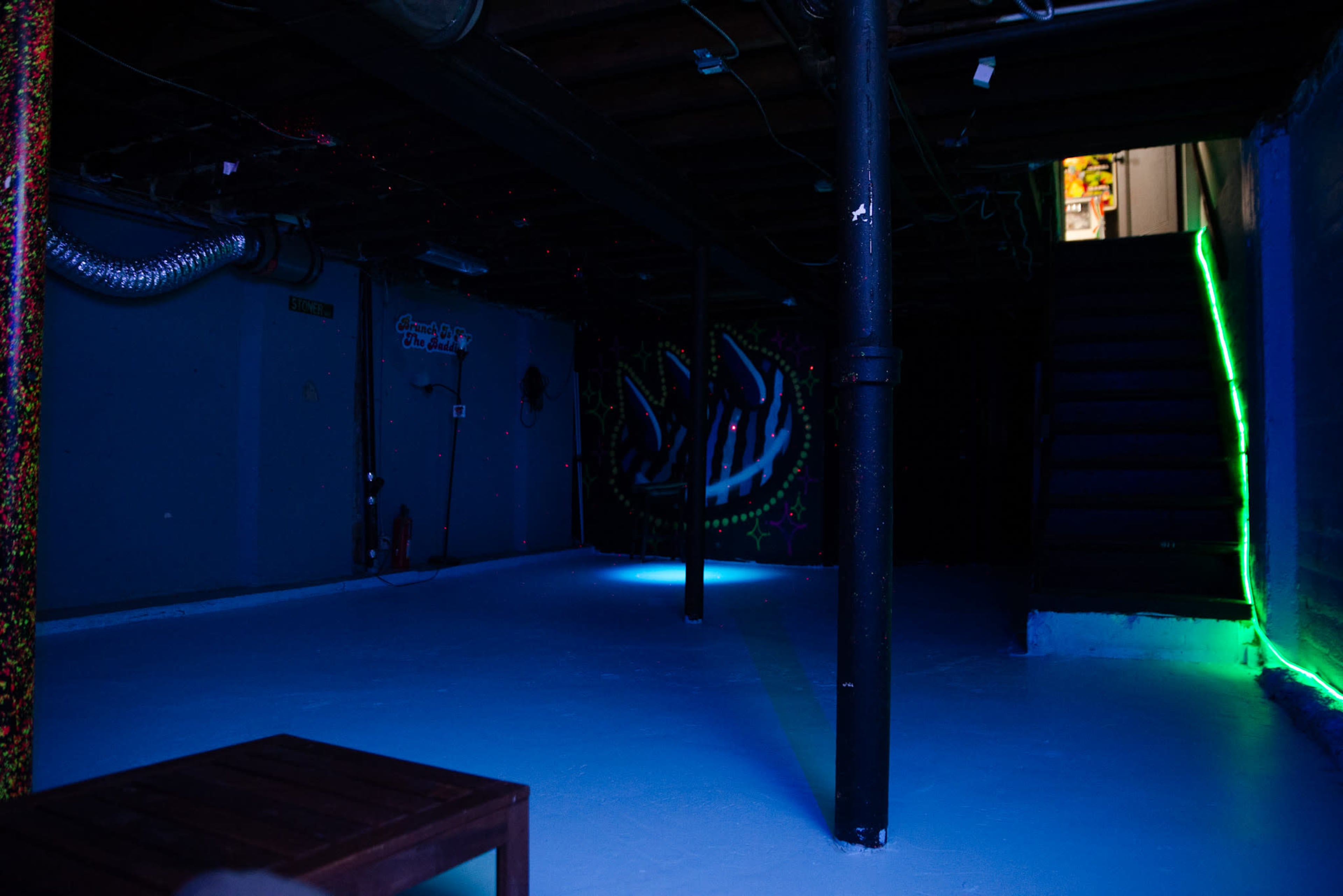 The image shows a dimly lit basement featuring a painted wall with a graphic design, a staircase leading to an upper level, and a small wooden table in the foreground.