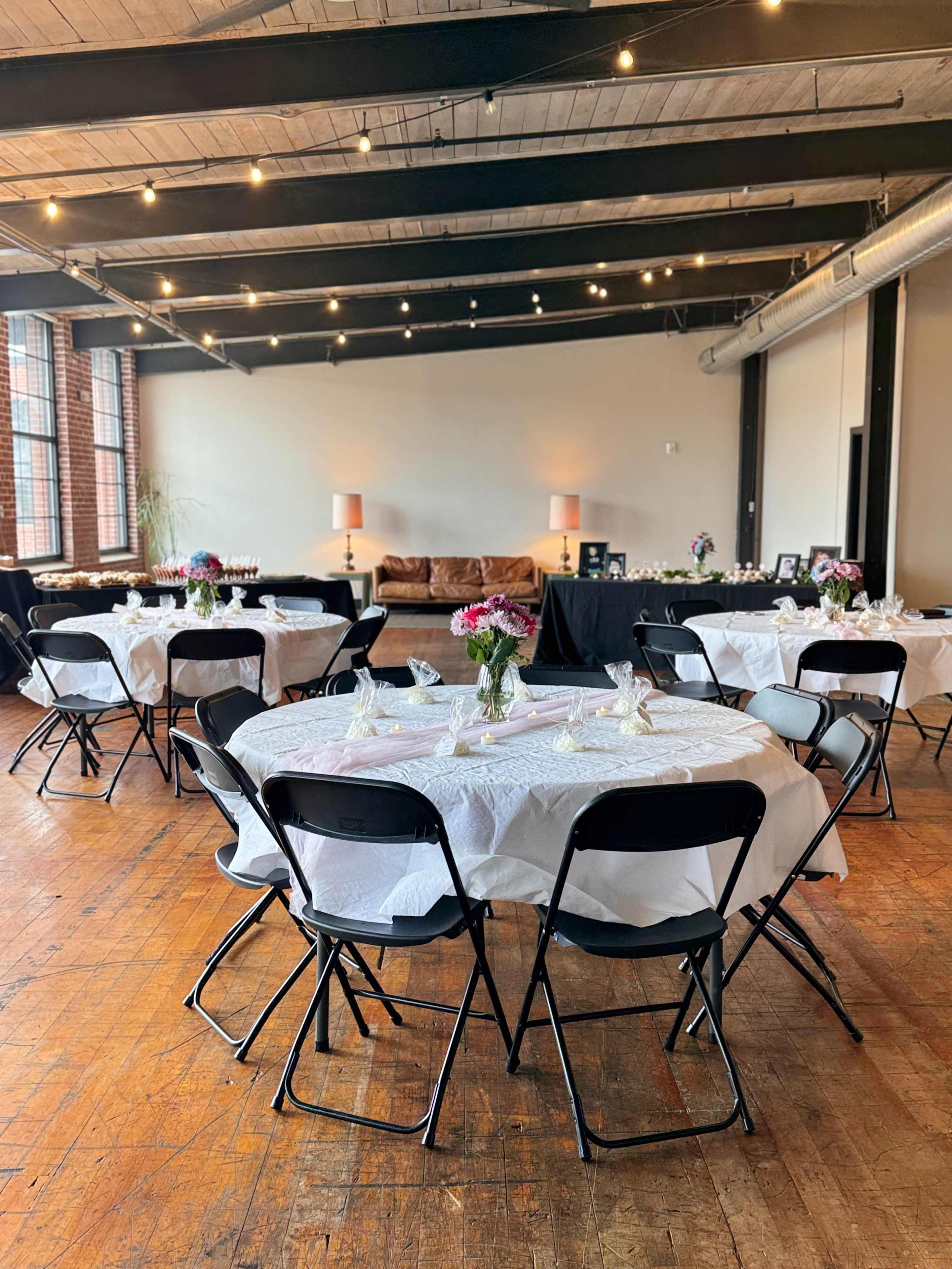 The image showcases a spacious event room set up with round tables covered in white tablecloths, each adorned with flowers and placed against a backdrop of exposed wooden beams and large windows.