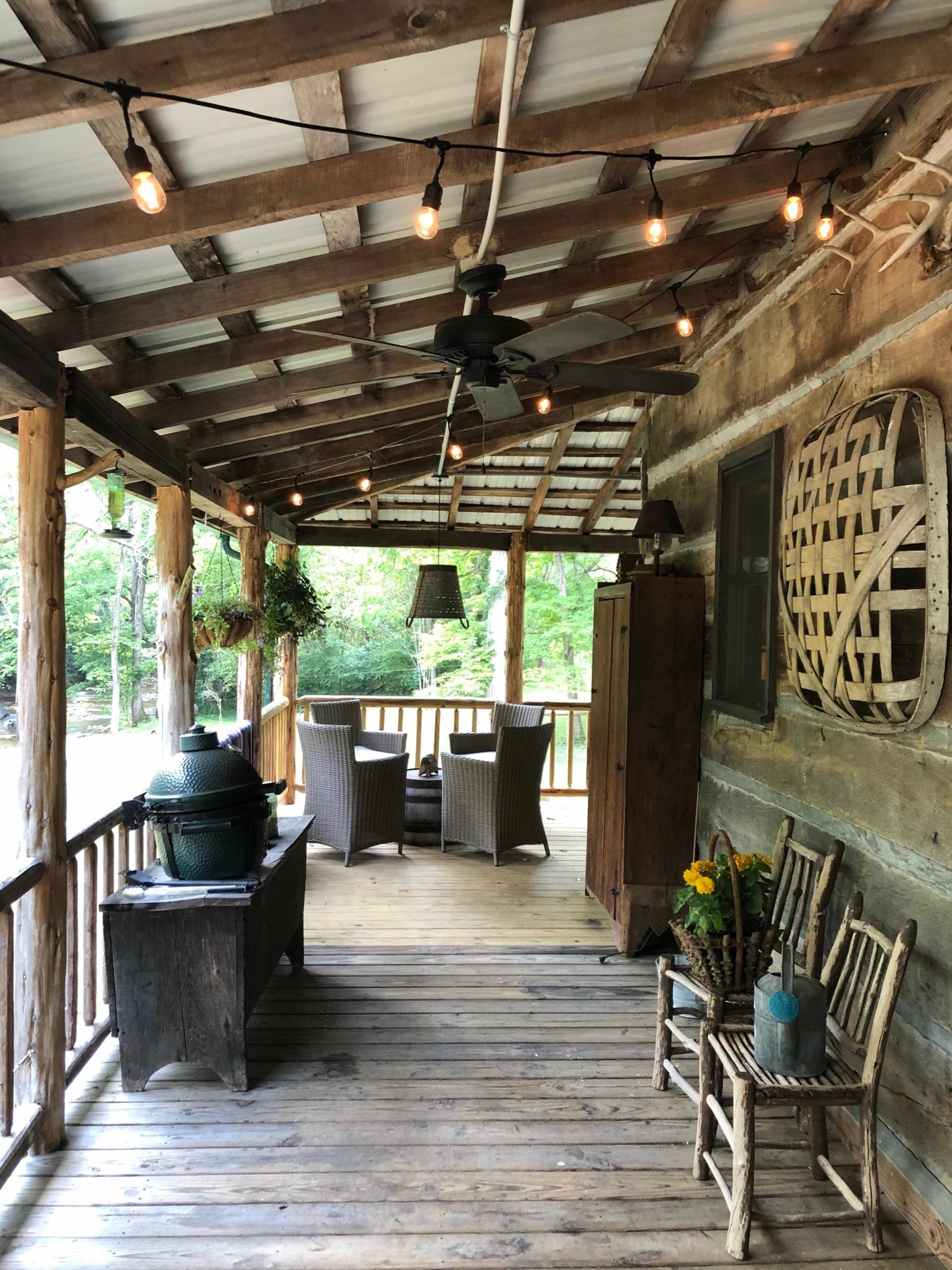 A rustic porch with wooden beams, a ceiling fan, and string lights, furnished with wicker chairs and a table, surrounded by greenery.