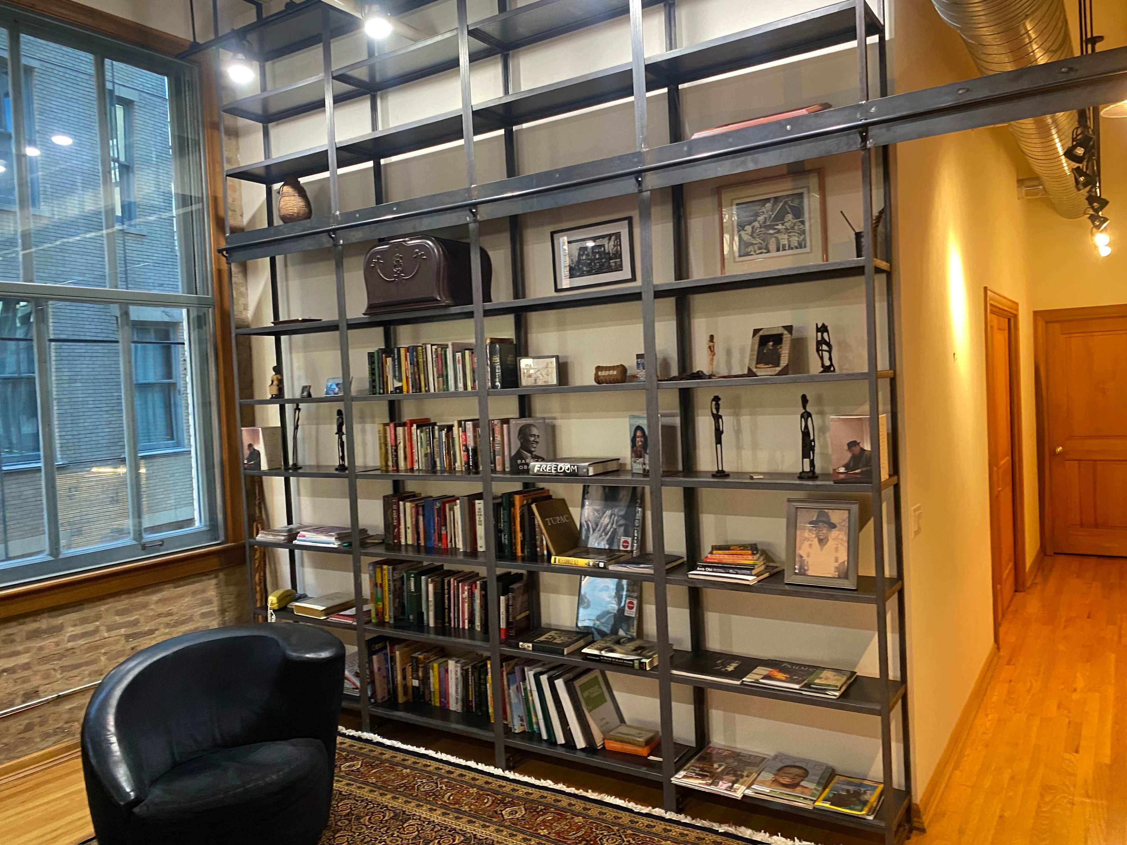 A modern room with a tall black shelving unit filled with books and decorative items, alongside a black armchair and large windows letting in natural light.