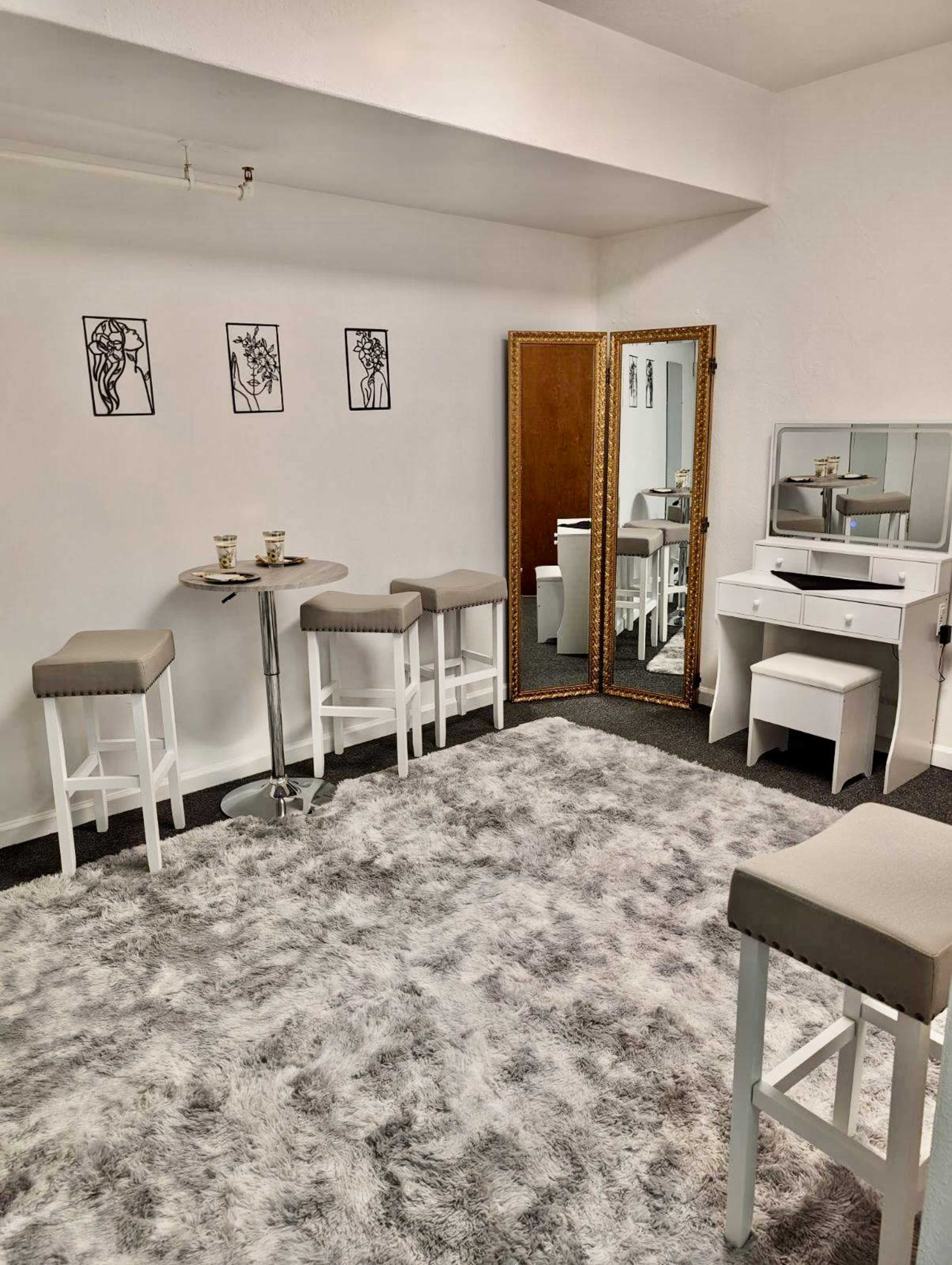 The image features a minimalist room with a plush gray carpet, white furniture including stools and a vanity, and three framed artworks on the walls.