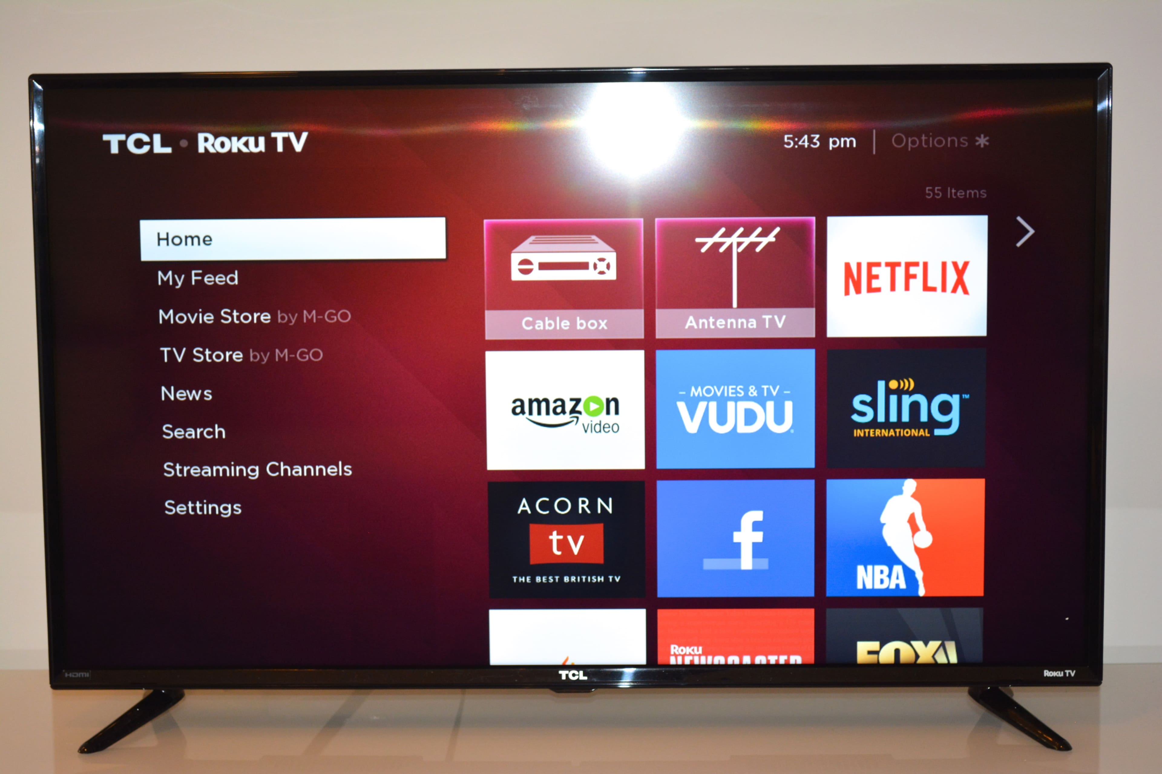 The image shows a TCL Roku TV displaying a menu screen with options for streaming services and settings.