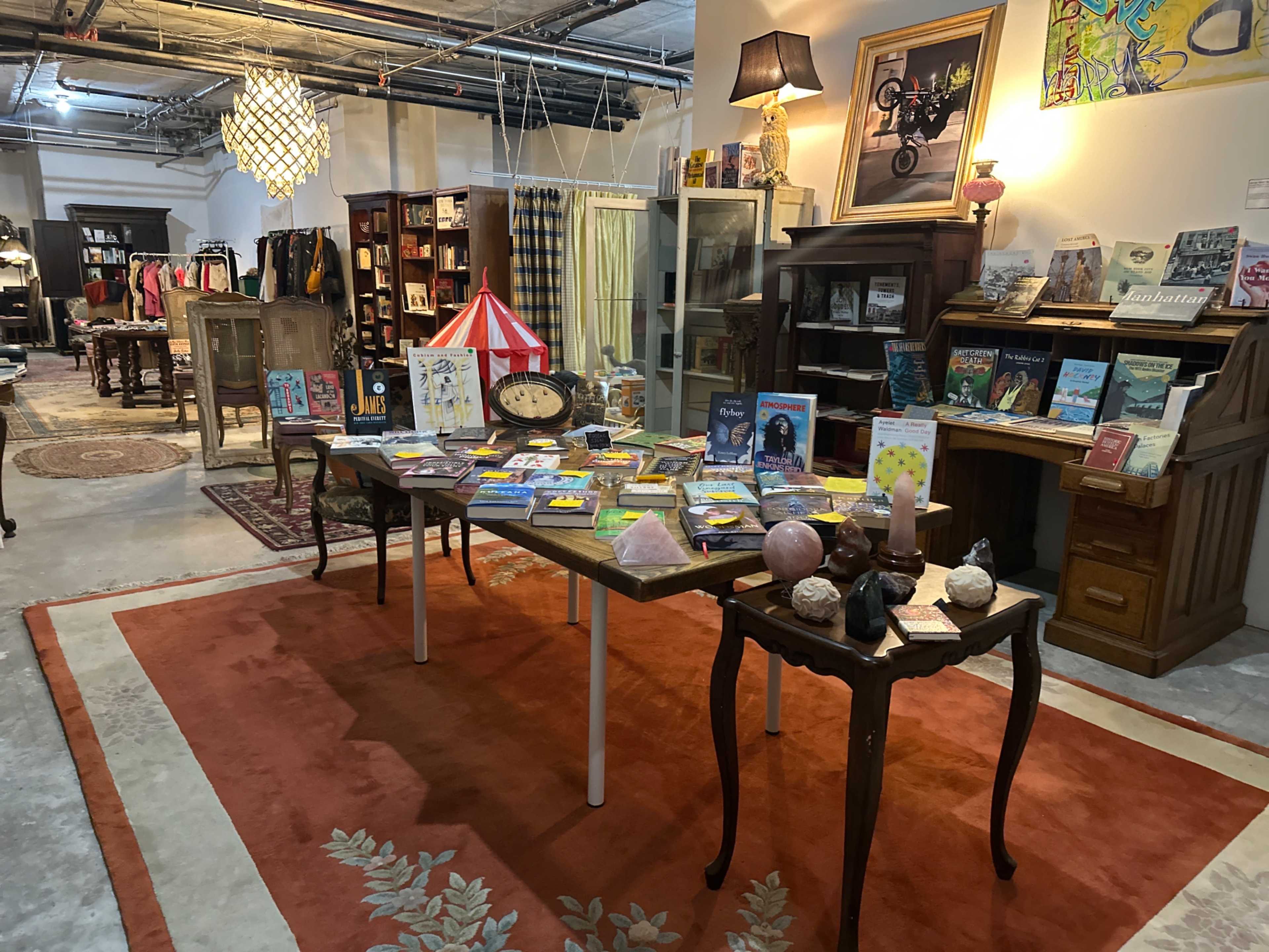 The image shows a cozy, well-lit shop interior with a table displaying books and decorative items, surrounded by vintage furniture and clothing racks.