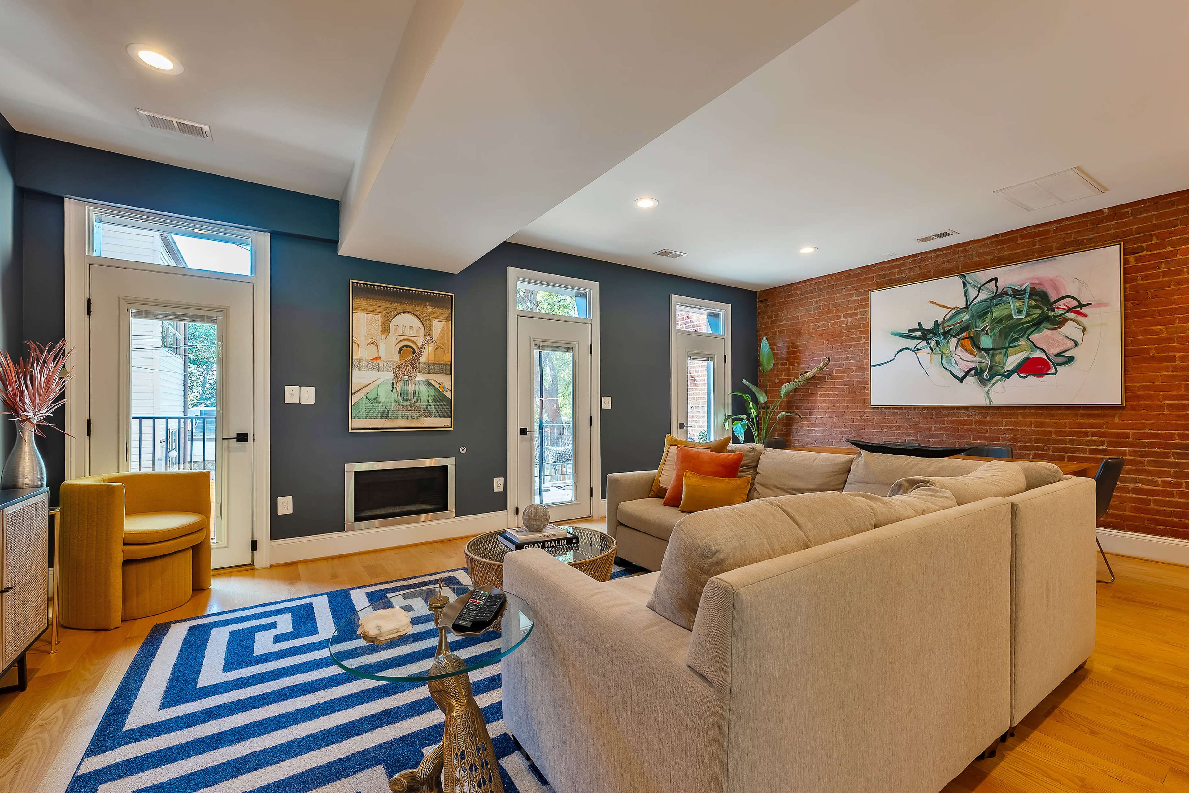 The living room features a modern sofa, a geometric rug, art on the walls, large windows, and a brick accent wall.