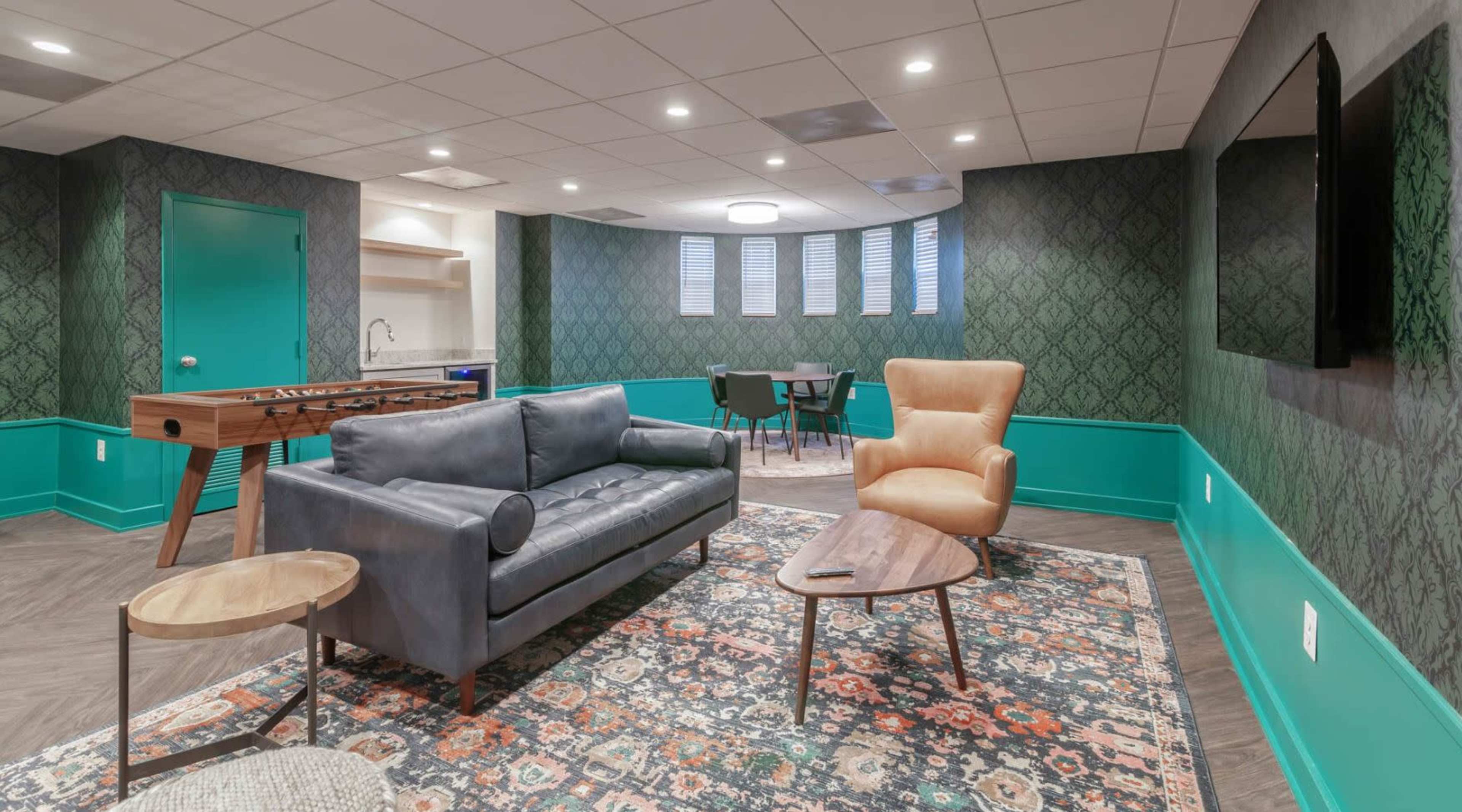 A modern lounge area featuring a dark couch, a light brown armchair, a coffee table, and a foosball table, all set against patterned wallpaper and teal accents.