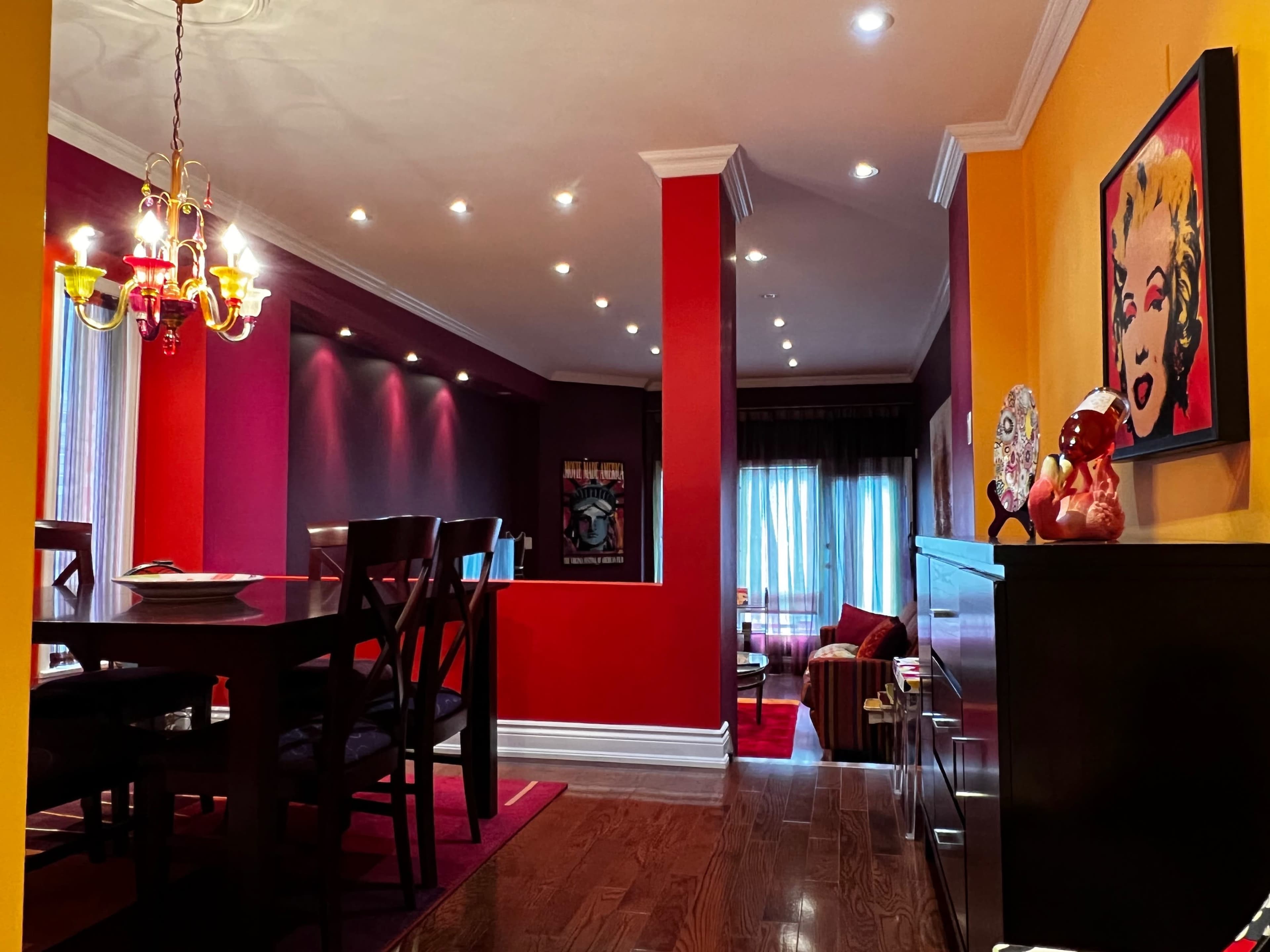 The room features a dining area with a dark wooden table and chairs, separated from a brightly colored living area by an open passage adorned with vibrant walls.