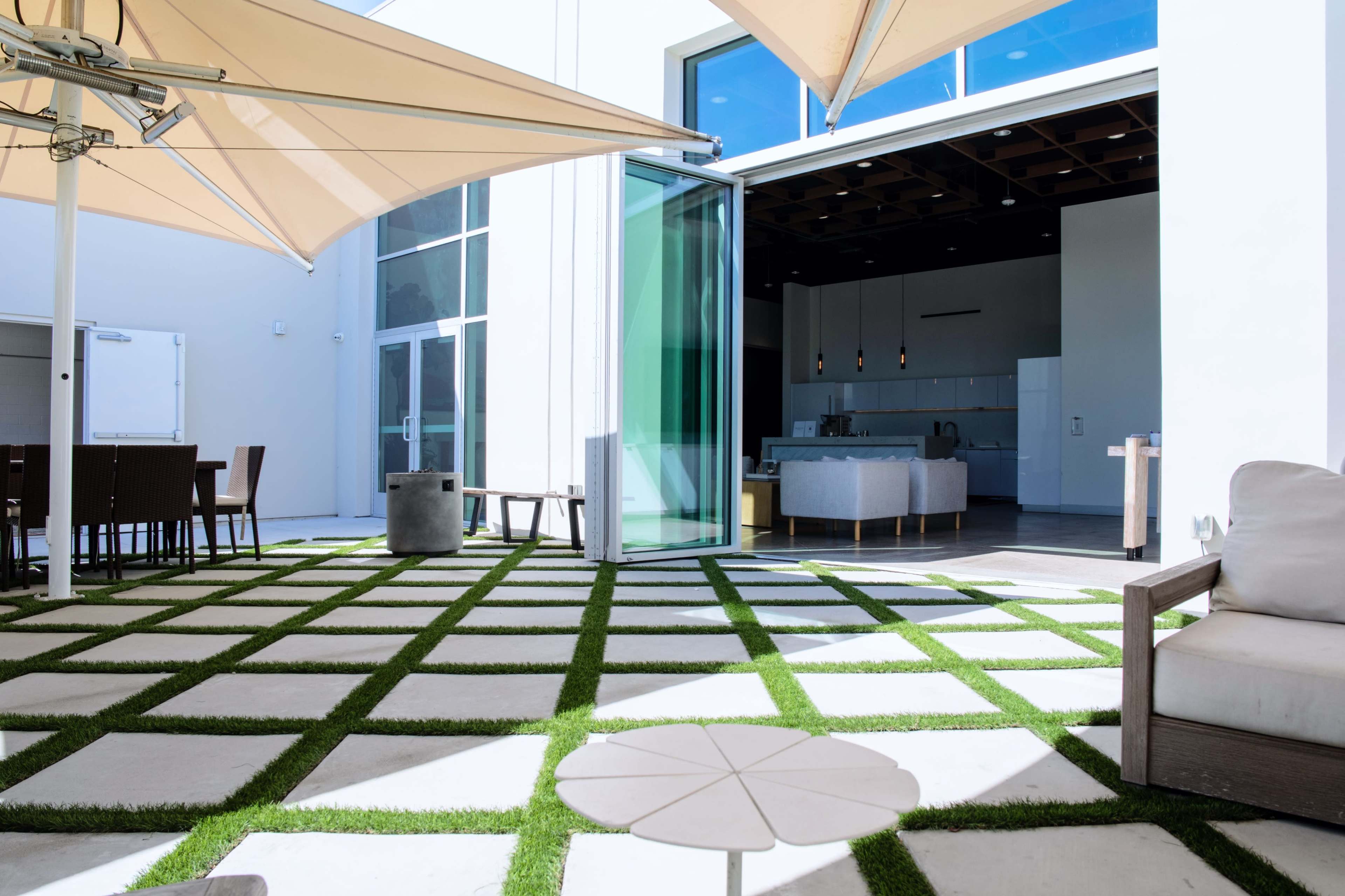 An outdoor patio features a grid of stone slabs surrounded by green grass, leading to an open kitchen area with large glass doors.