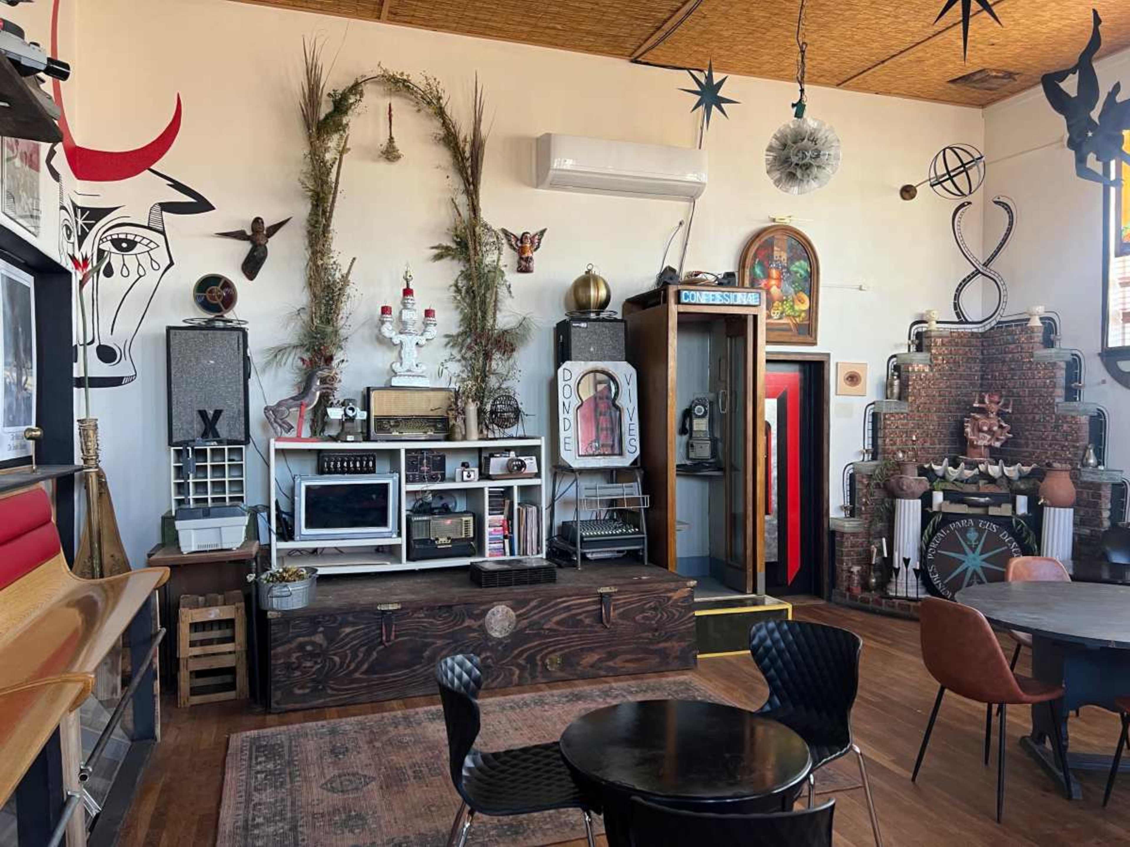 The image shows a retro-style interior with eclectic decor, including vintage appliances, artwork, and various ornamental objects, centered around a wooden table and chairs.