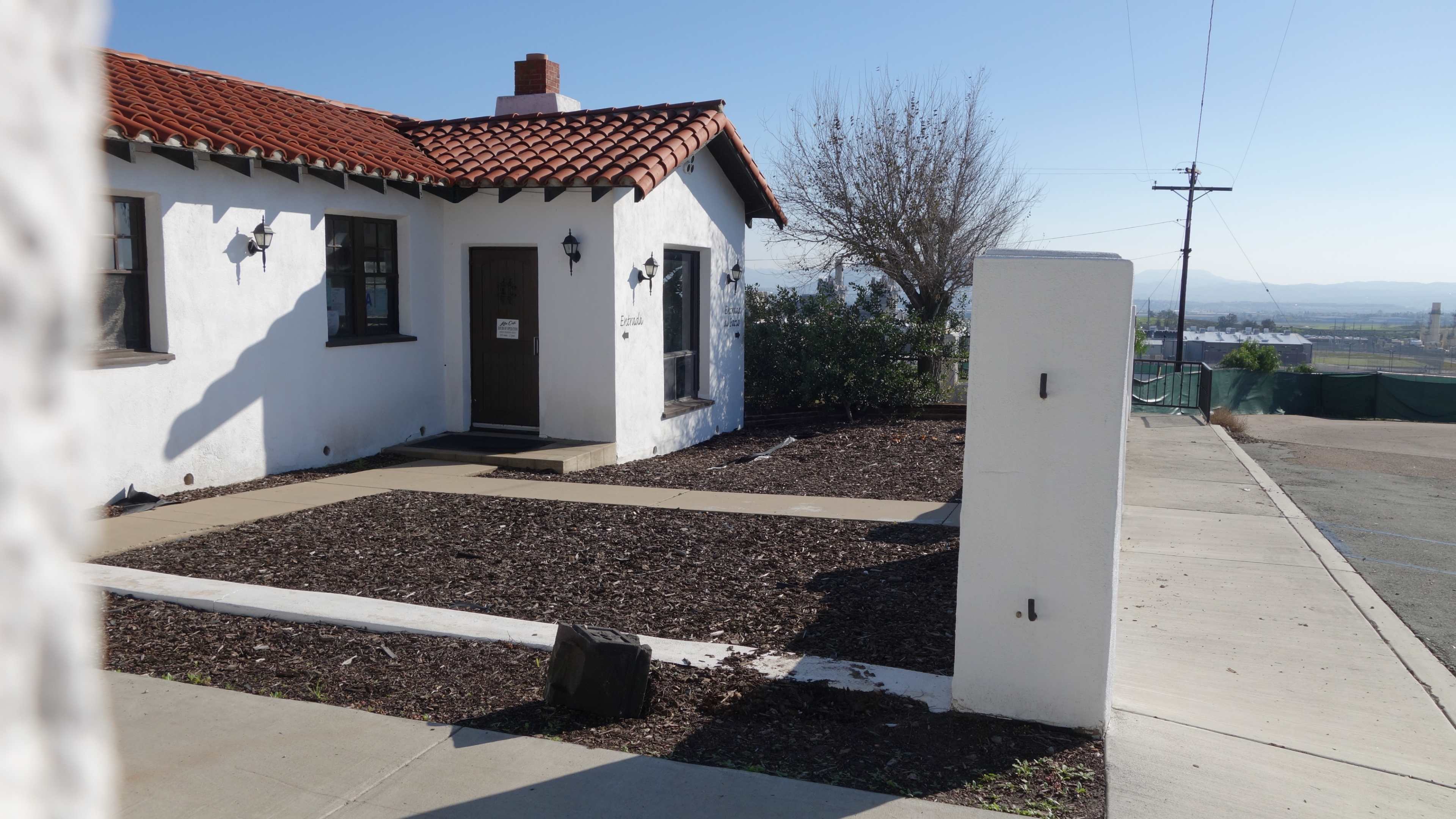 A small, white stucco house with a red-tiled roof, situated on a landscaped yard with mulch and a concrete pathway.