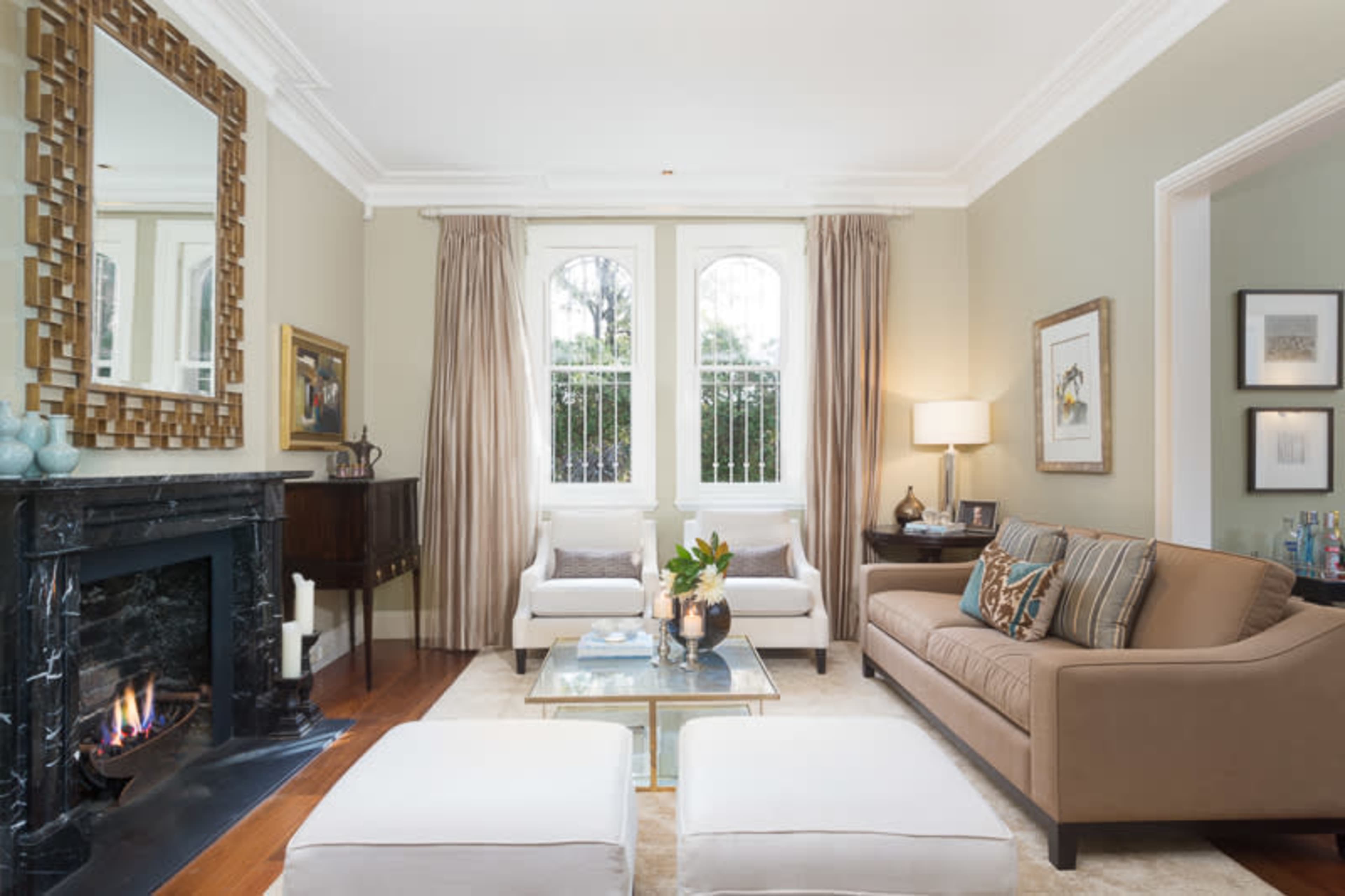 A living room with a brown sofa, two white chairs, a glass coffee table, and a fireplace, along with large windows covered by curtains.